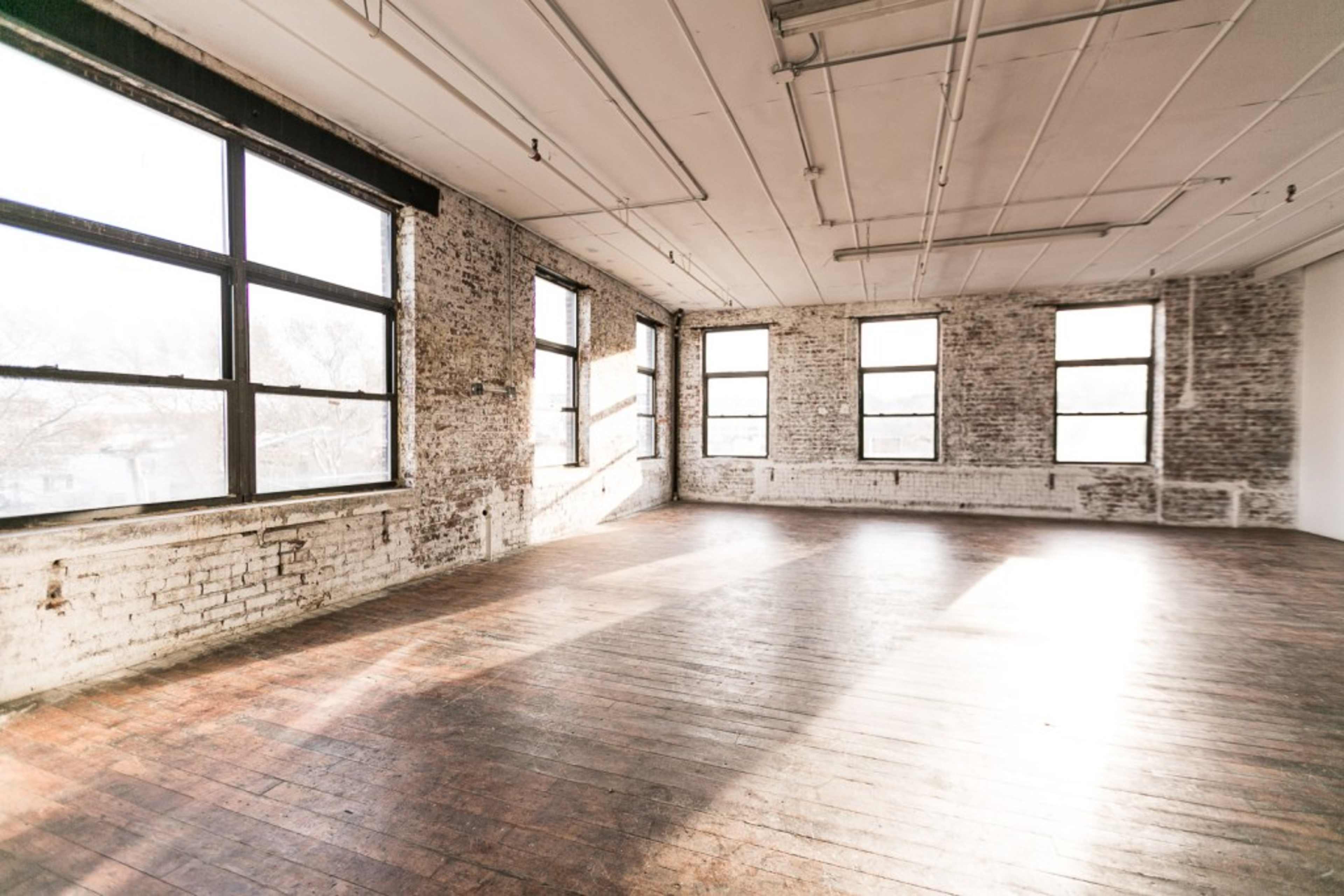 An empty room with large windows and exposed brick walls, featuring wooden floors illuminated by natural light.