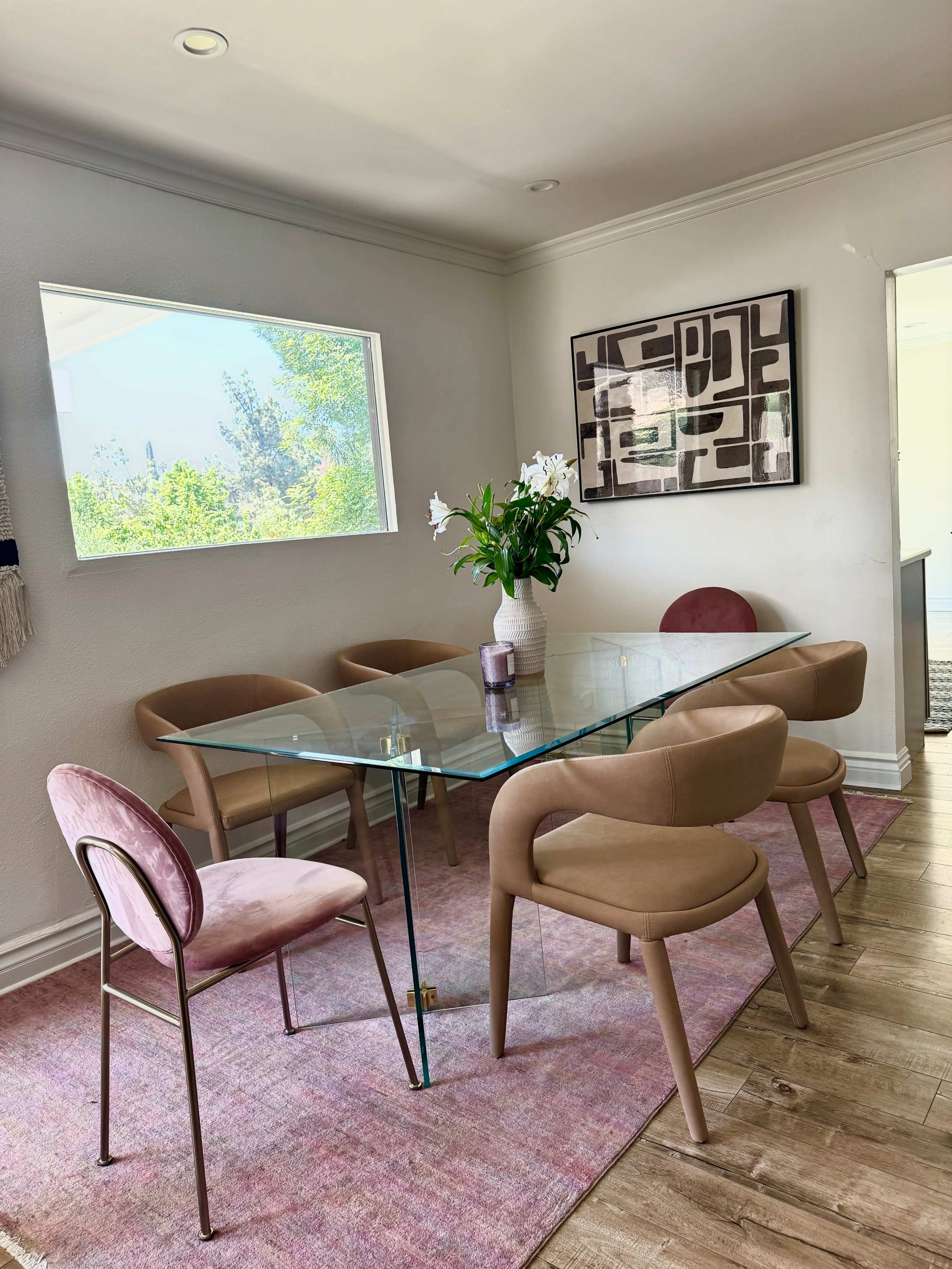 A glass dining table is set in a well-lit room, surrounded by pink upholstered chairs, with a vase of white flowers at the center and a patterned artwork on the wall.