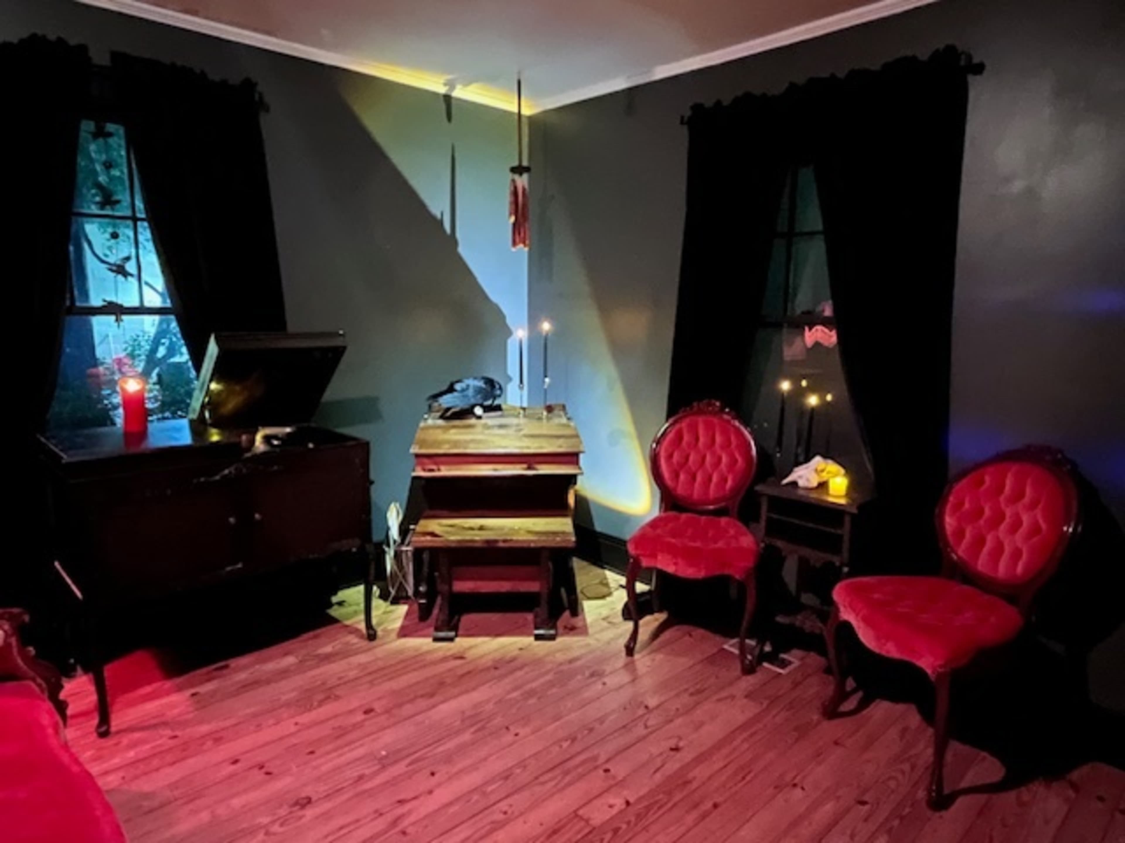 The image shows a dimly lit room with dark walls, a wooden floor, a vintage wooden chest, two red upholstered chairs, and candles on a small table.