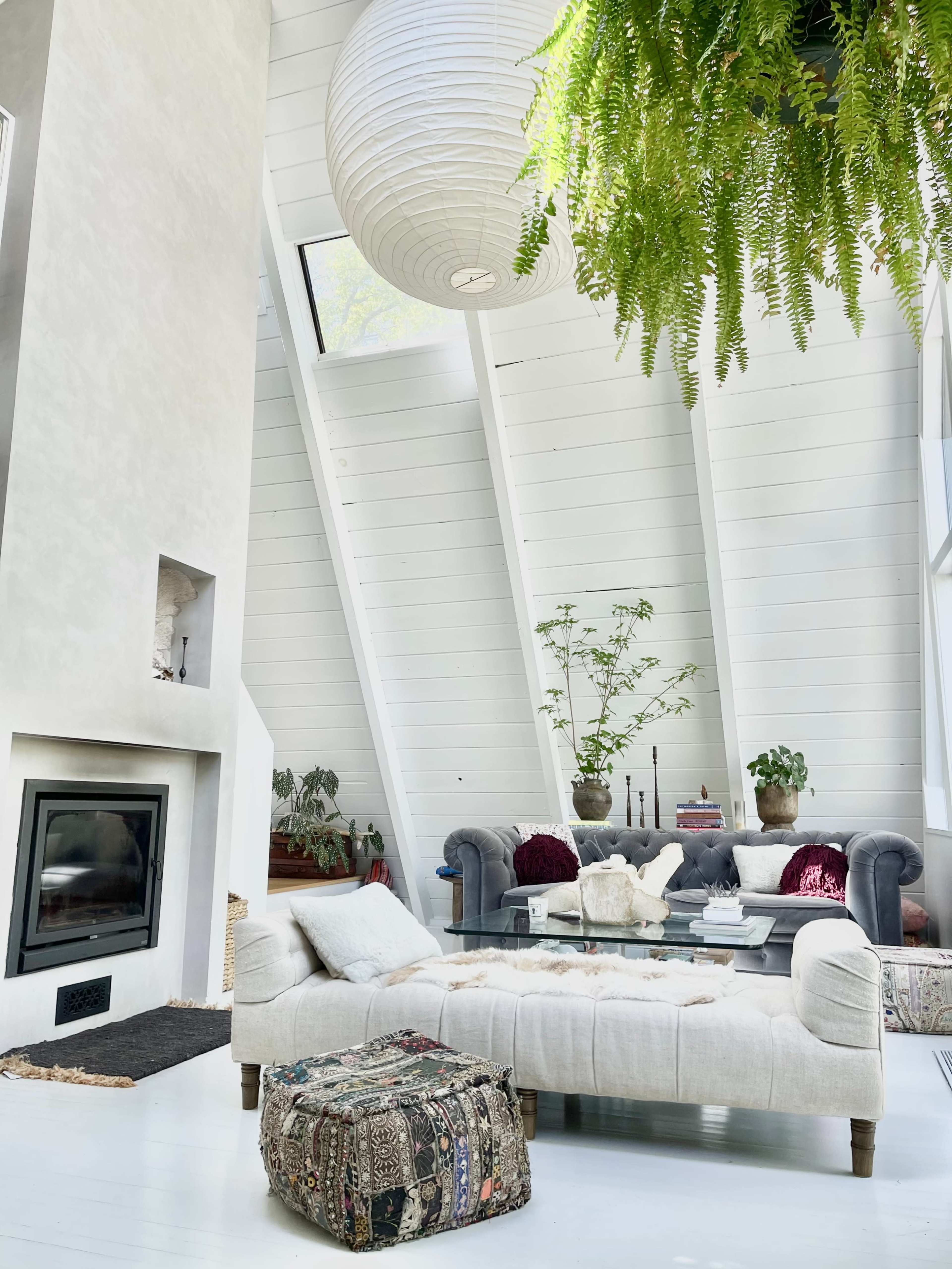 The living room features a modern design with large windows, a cozy sofa, a glass coffee table, and hanging plants.