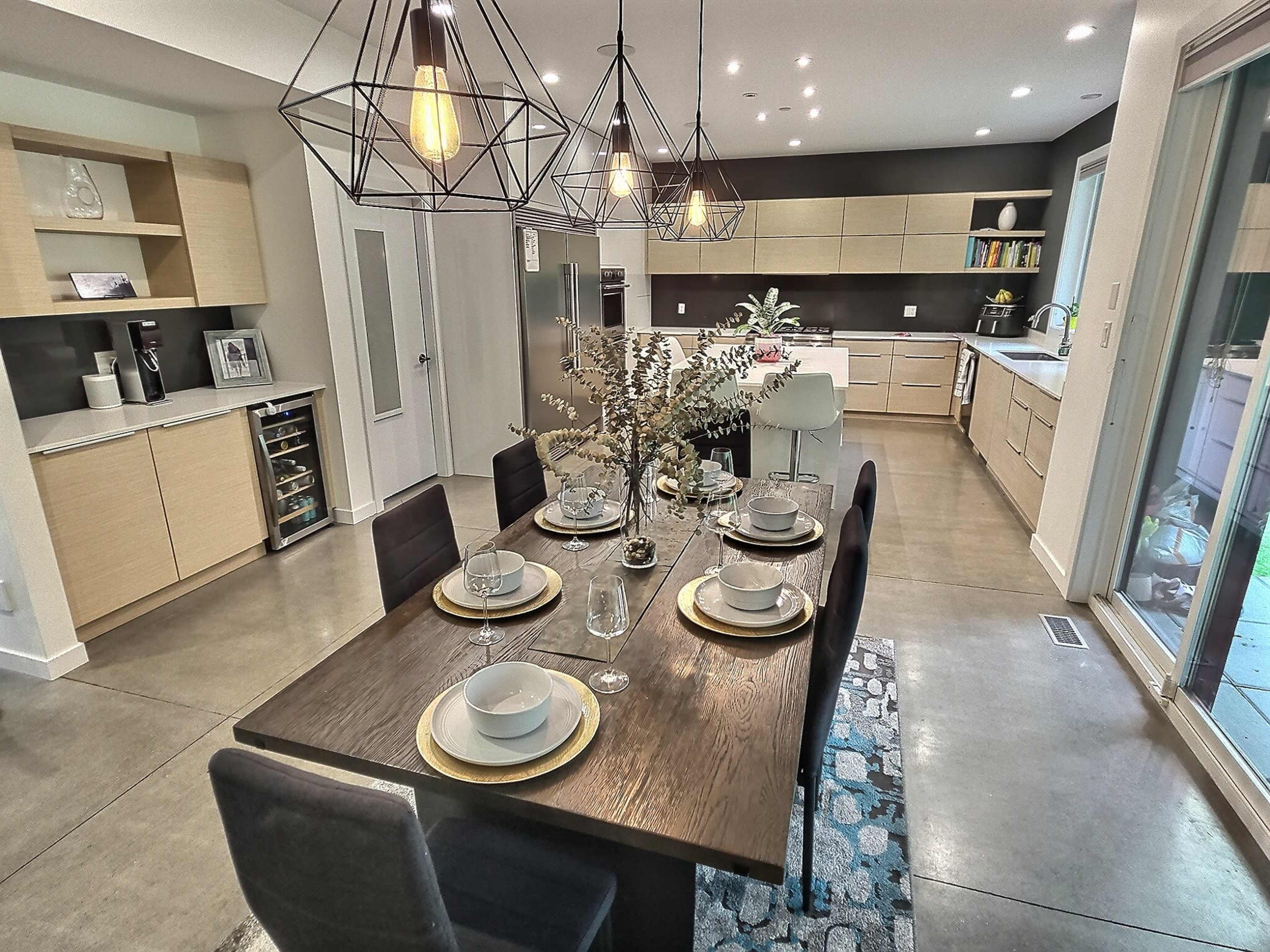 A modern dining area features a wooden table set for a meal, surrounded by black chairs, with pendant lights overhead and a view of a kitchen in the background.