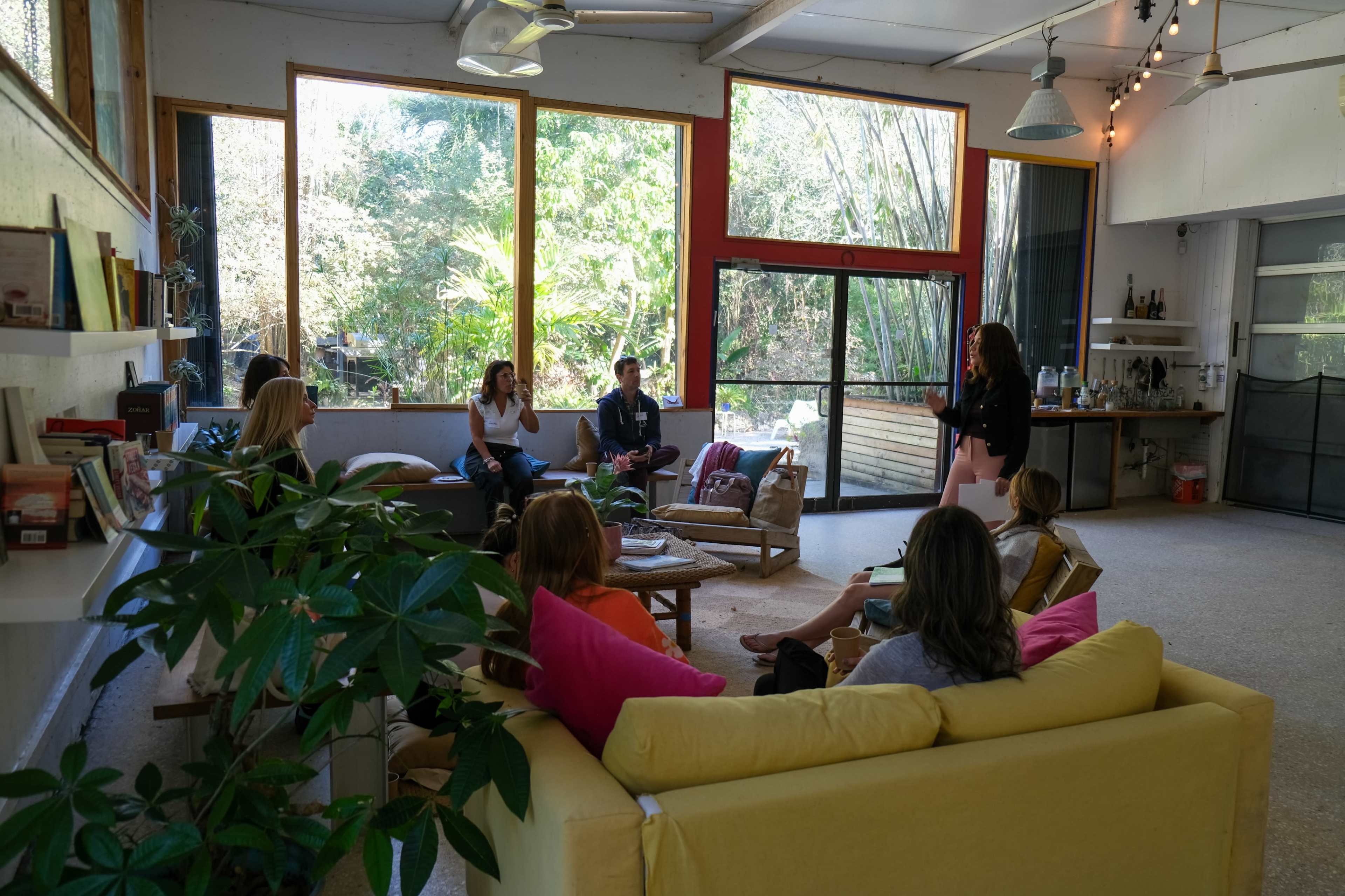 A group of eight people sits in a spacious room with large windows, discussing while surrounded by plants and modern decor.