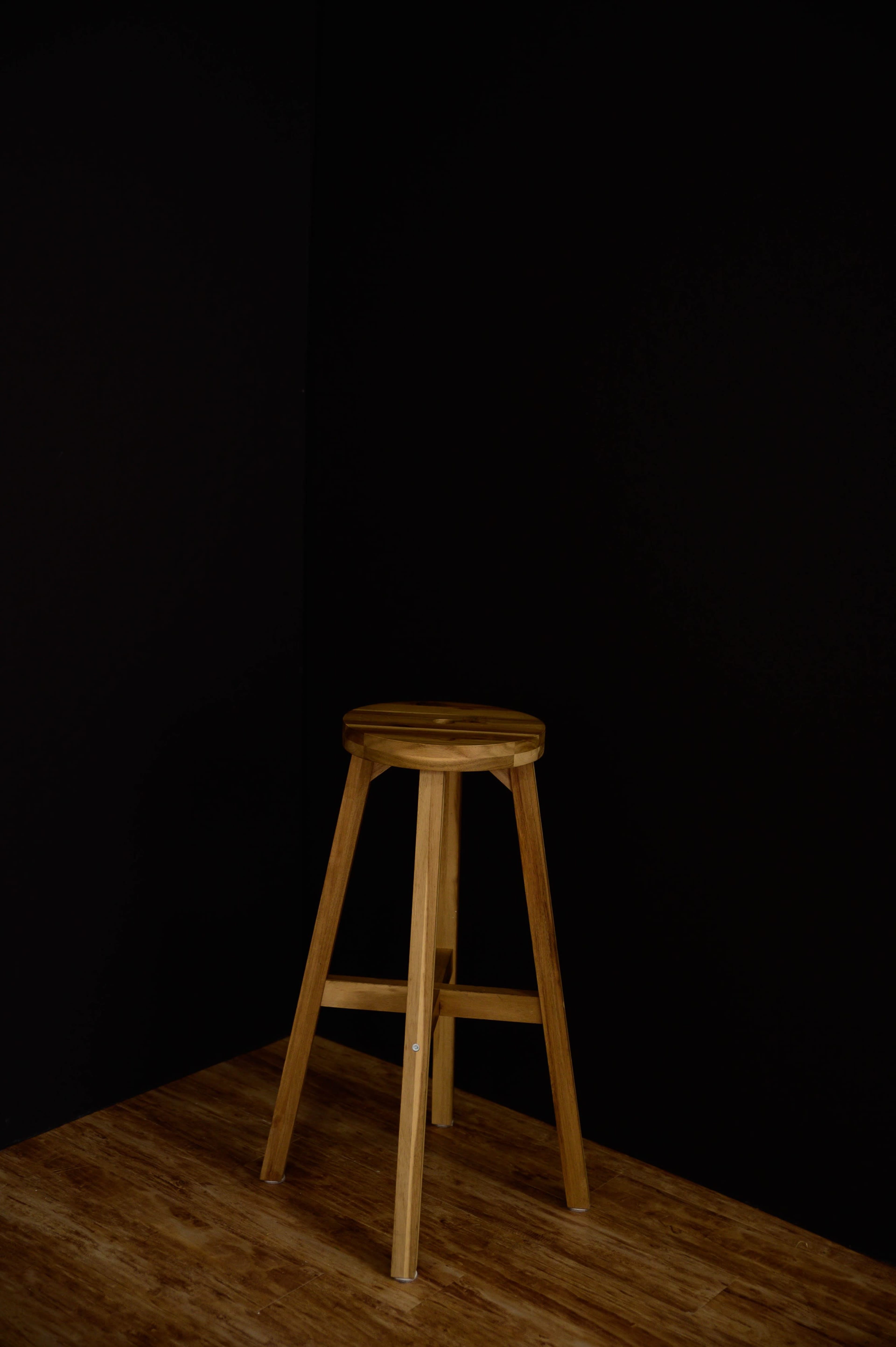 A wooden stool stands alone in a corner against a black wall.