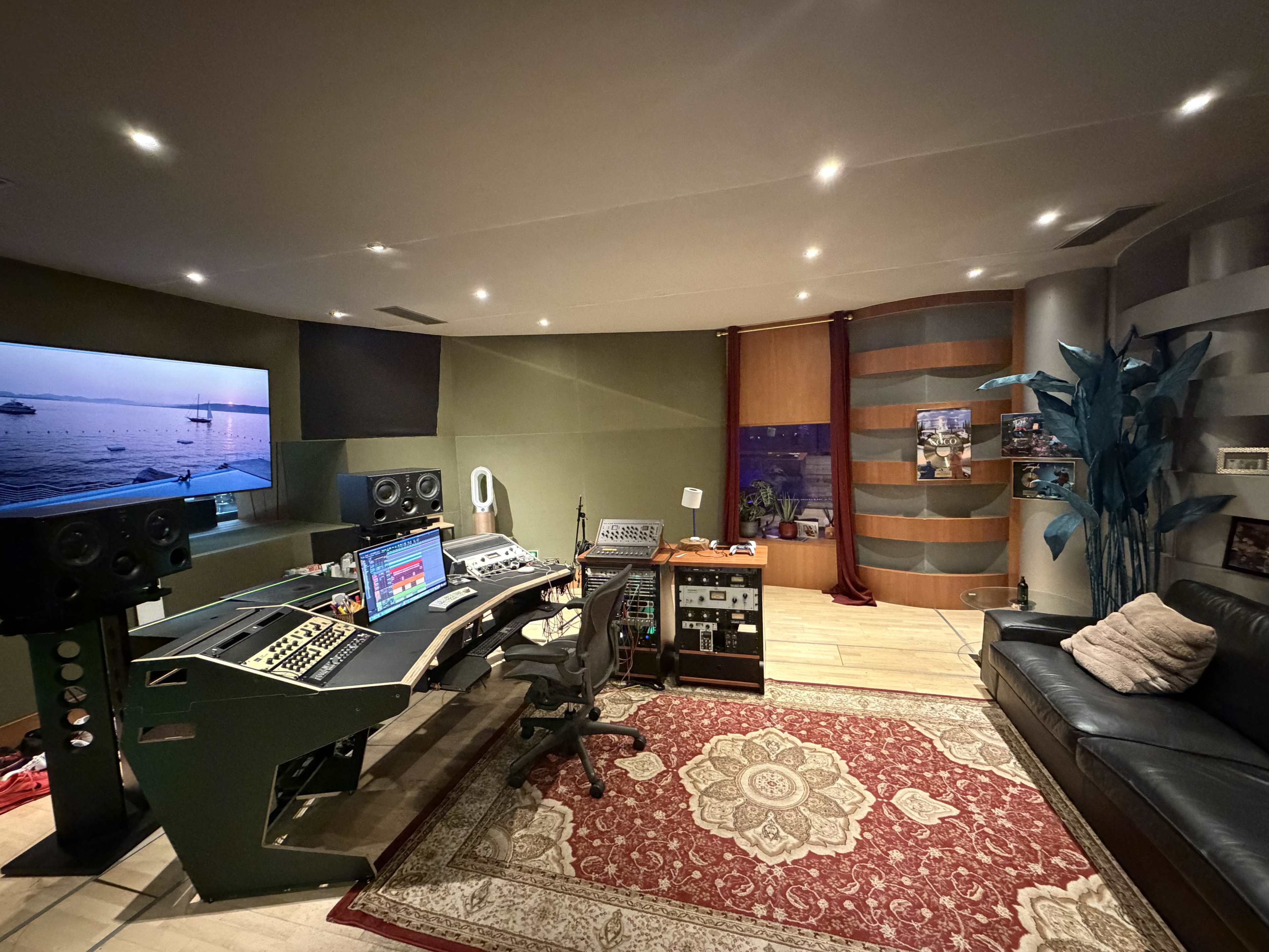 The image shows a recording studio featuring a central mixing console, large speakers, a black leather sofa, and a decorative area rug on the floor.