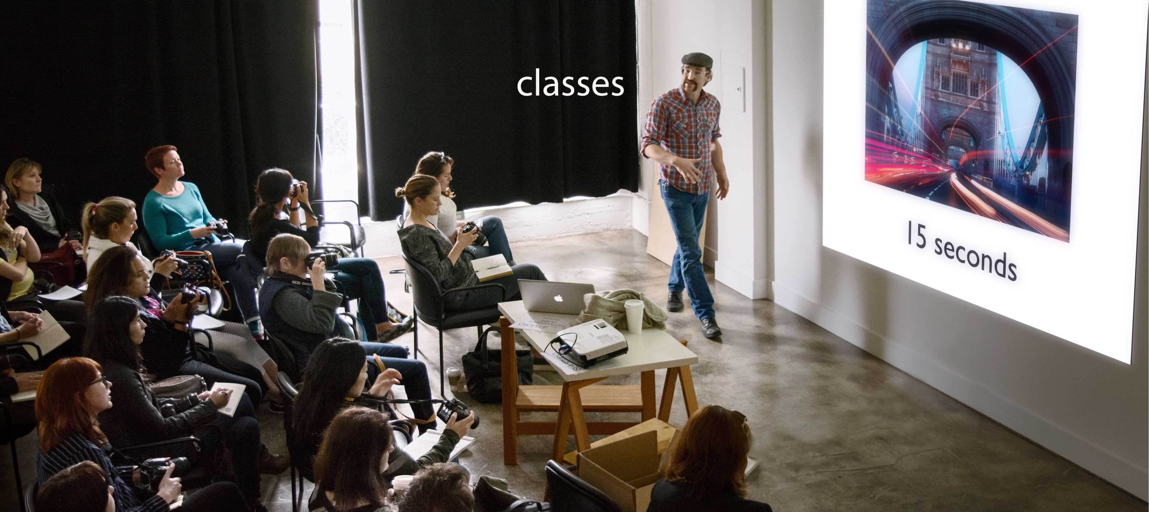 A photographer is giving a presentation to an audience seated in a studio setting, with a large projection displaying an image on the wall.