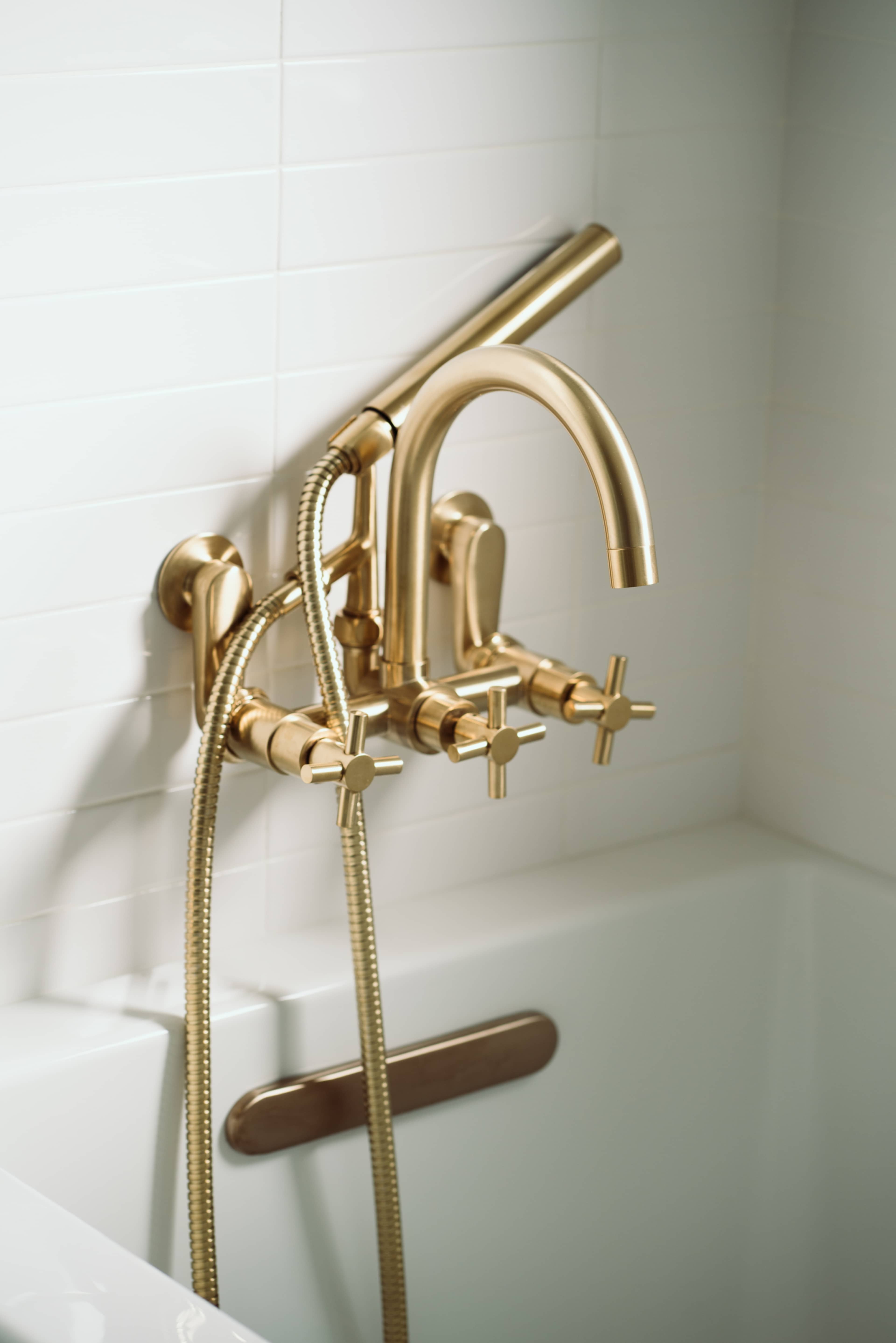 The image shows a gold bath faucet with a handheld shower attachment mounted on a white tiled wall.
