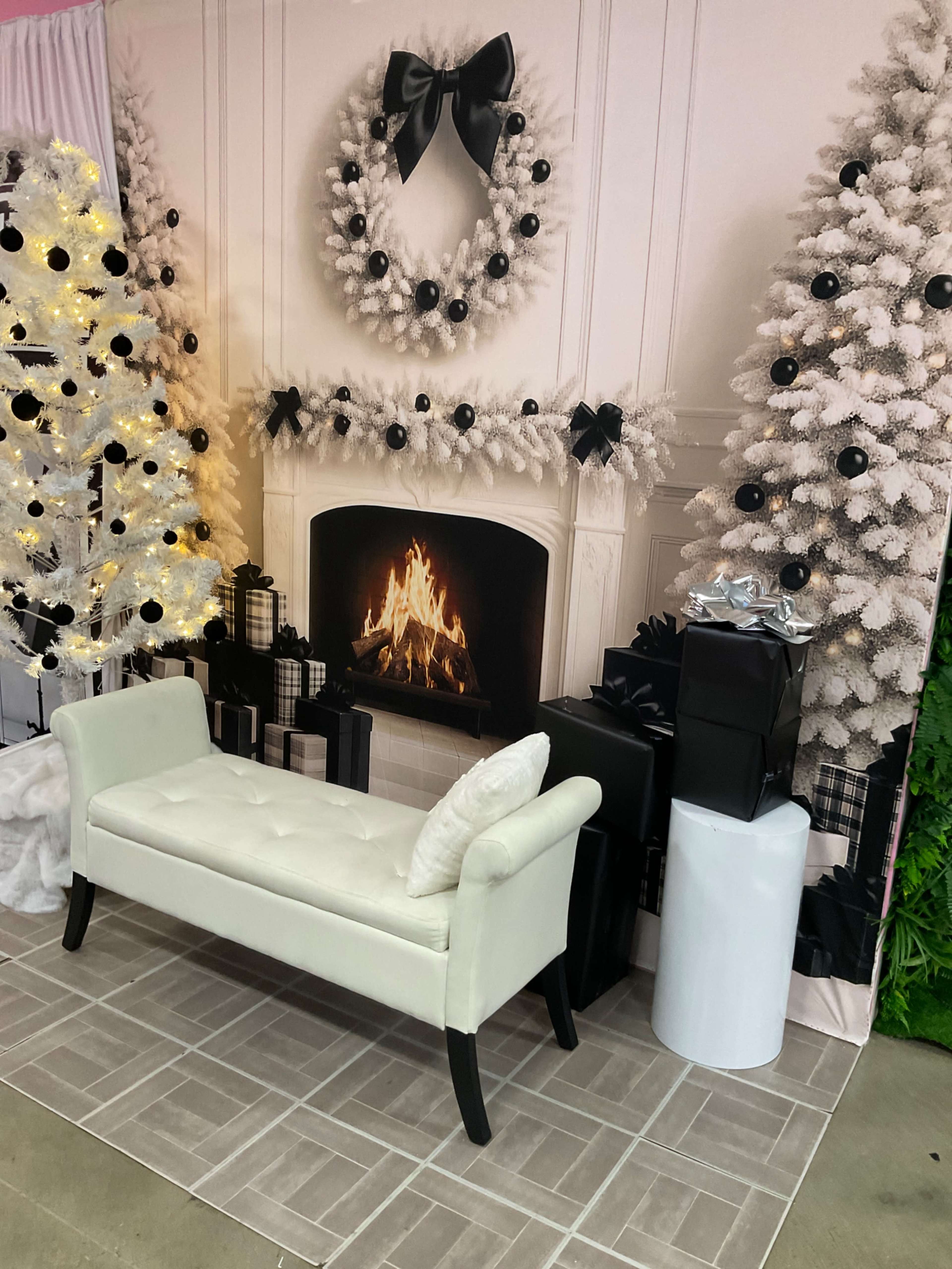 The image shows a festive display featuring a faux fireplace with a lit fire, surrounded by a white wreath and two decorated Christmas trees, alongside a white bench and wrapped presents.