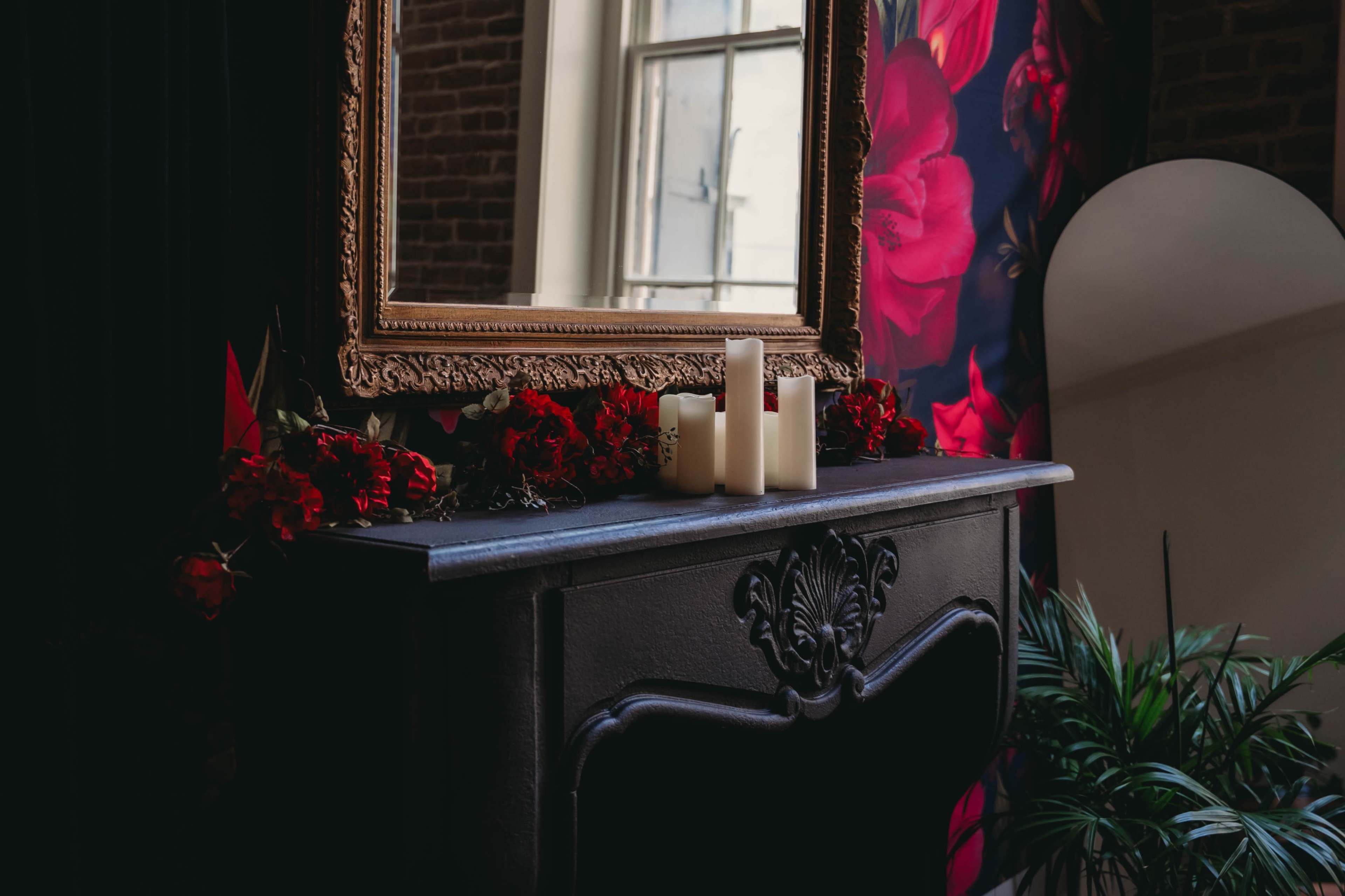 A dark wooden console table holds white candles and red flowers, reflecting a mirror against a floral-patterned wall.
