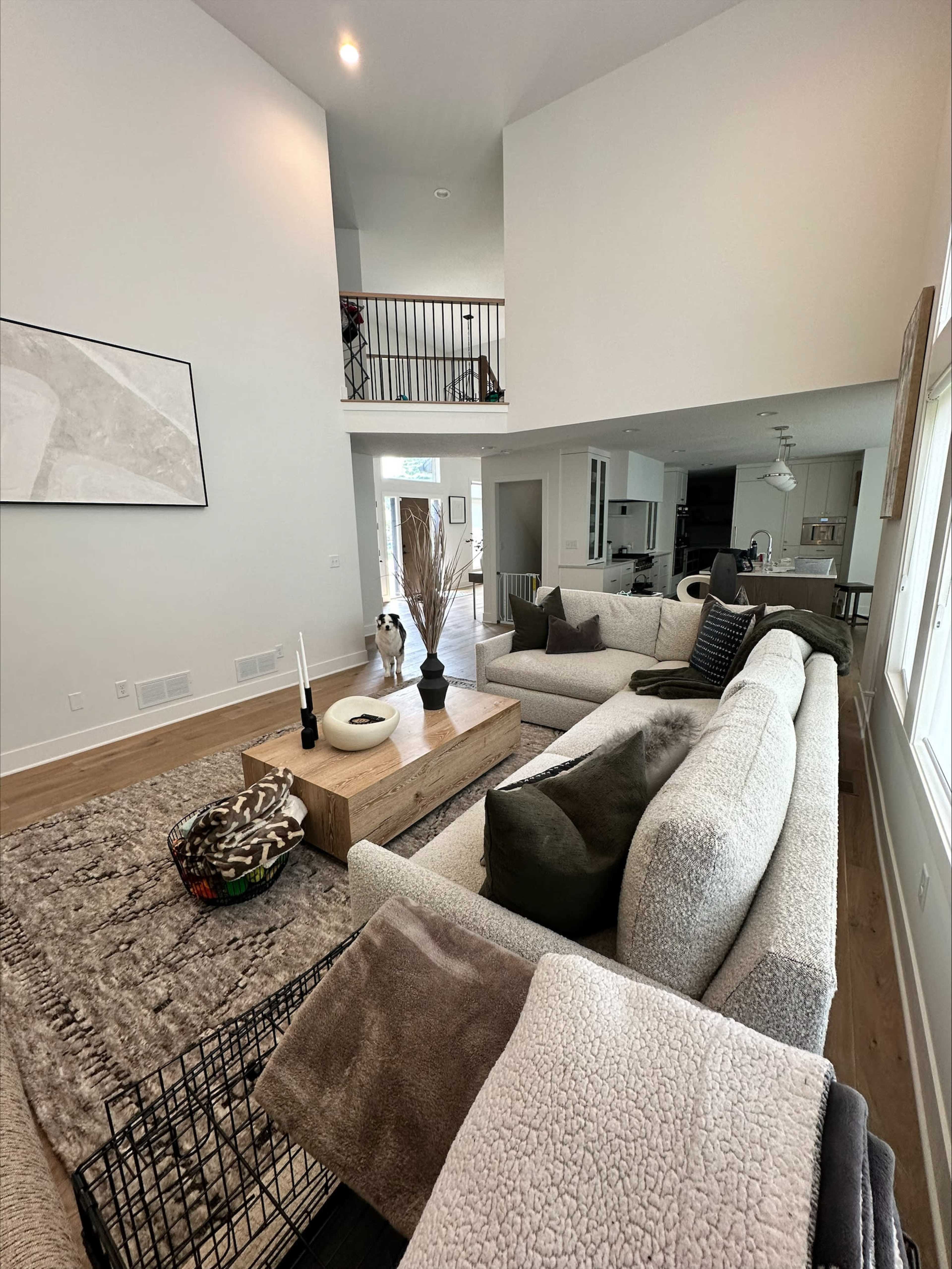 A spacious living room with a light-colored sectional sofa, a wooden coffee table, and a dog standing near a crate, with a staircase and kitchen area visible in the background.