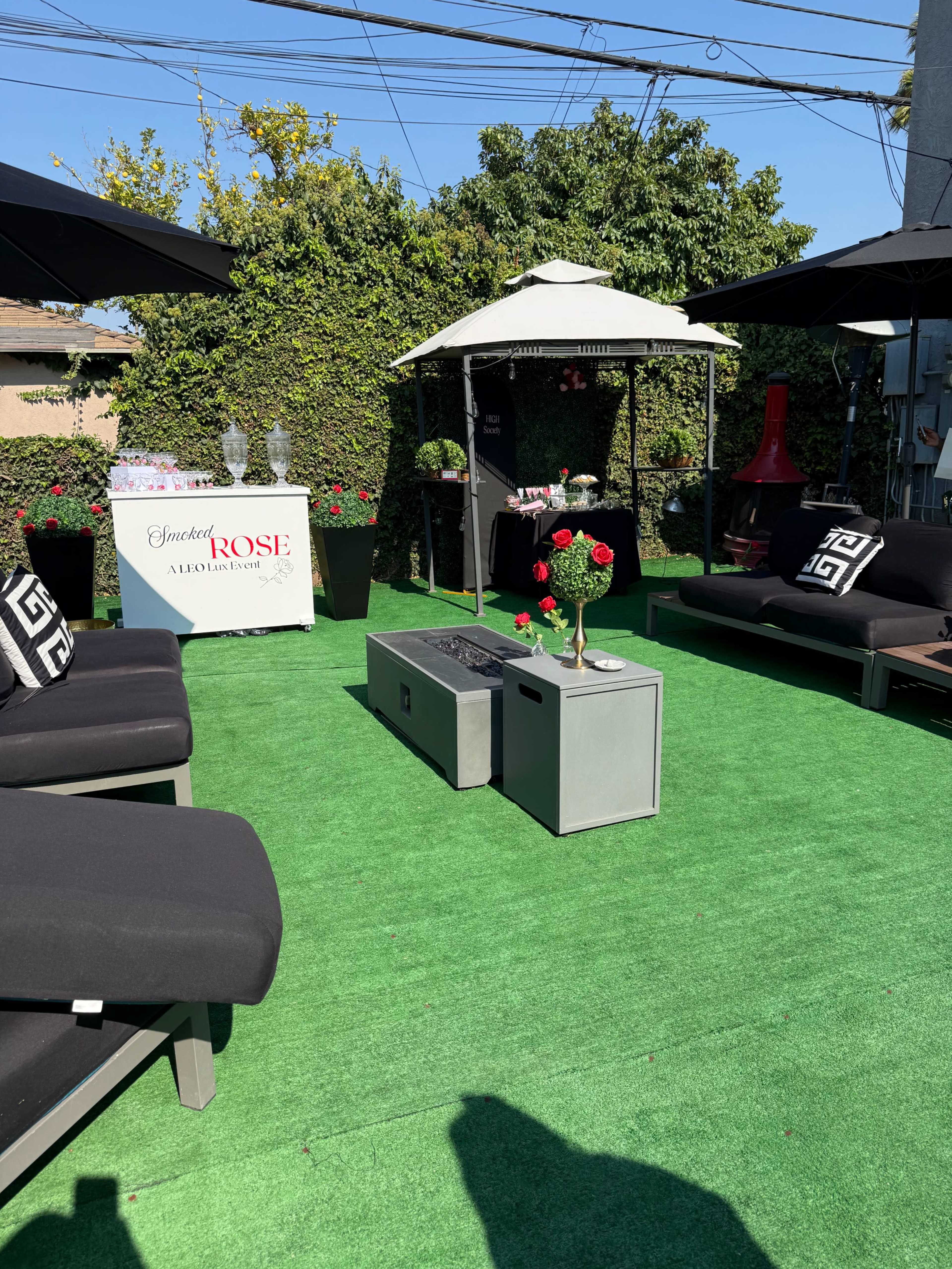 The image shows a well-arranged outdoor seating area with black lounge sofas, a central fire table, and a decorated kiosk under an umbrella, surrounded by greenery.