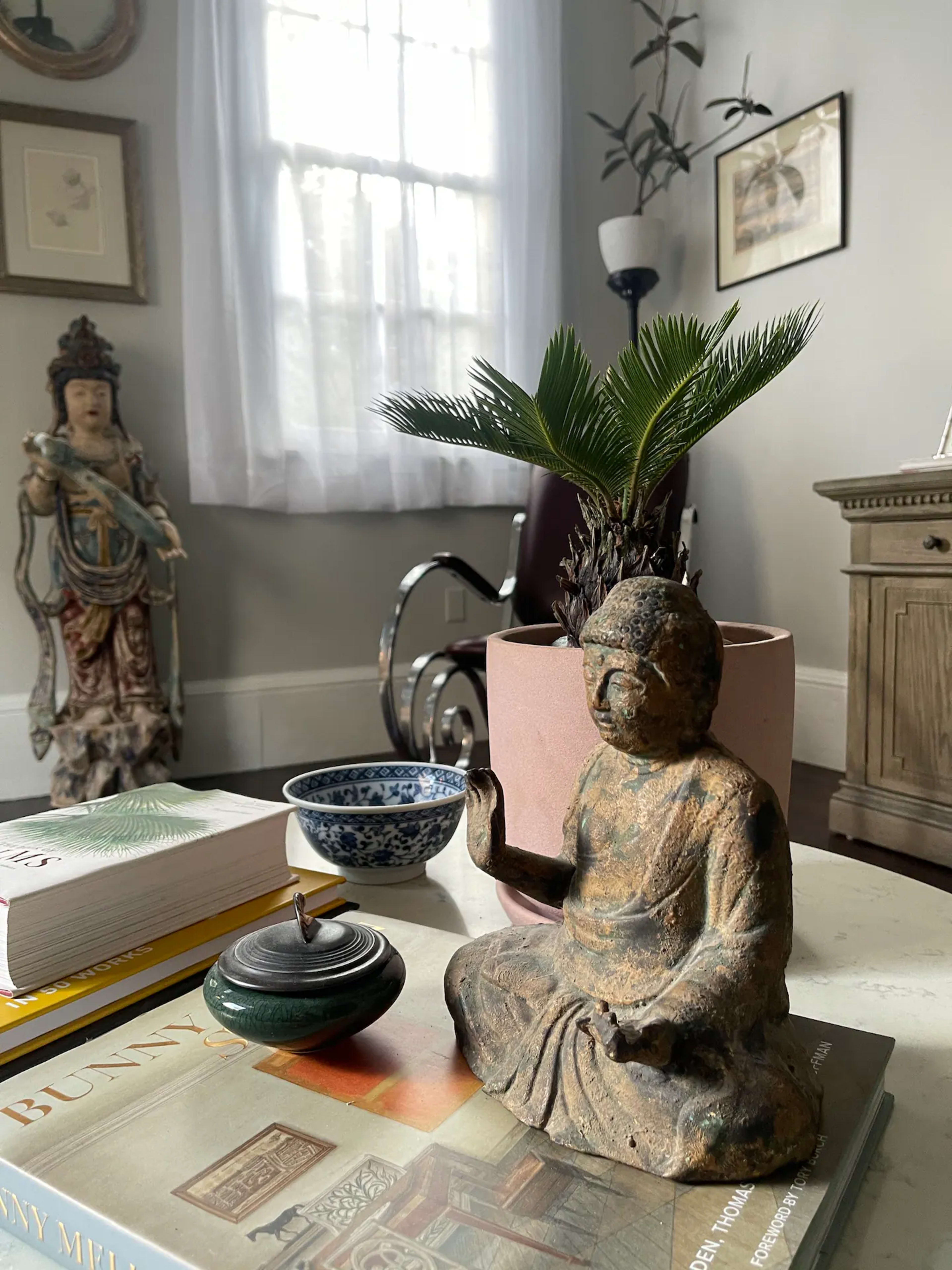 A small bronze Buddha statue sits on a coffee table alongside a potted plant, a bowl, and a candle, with sunlight streaming through a nearby window.
