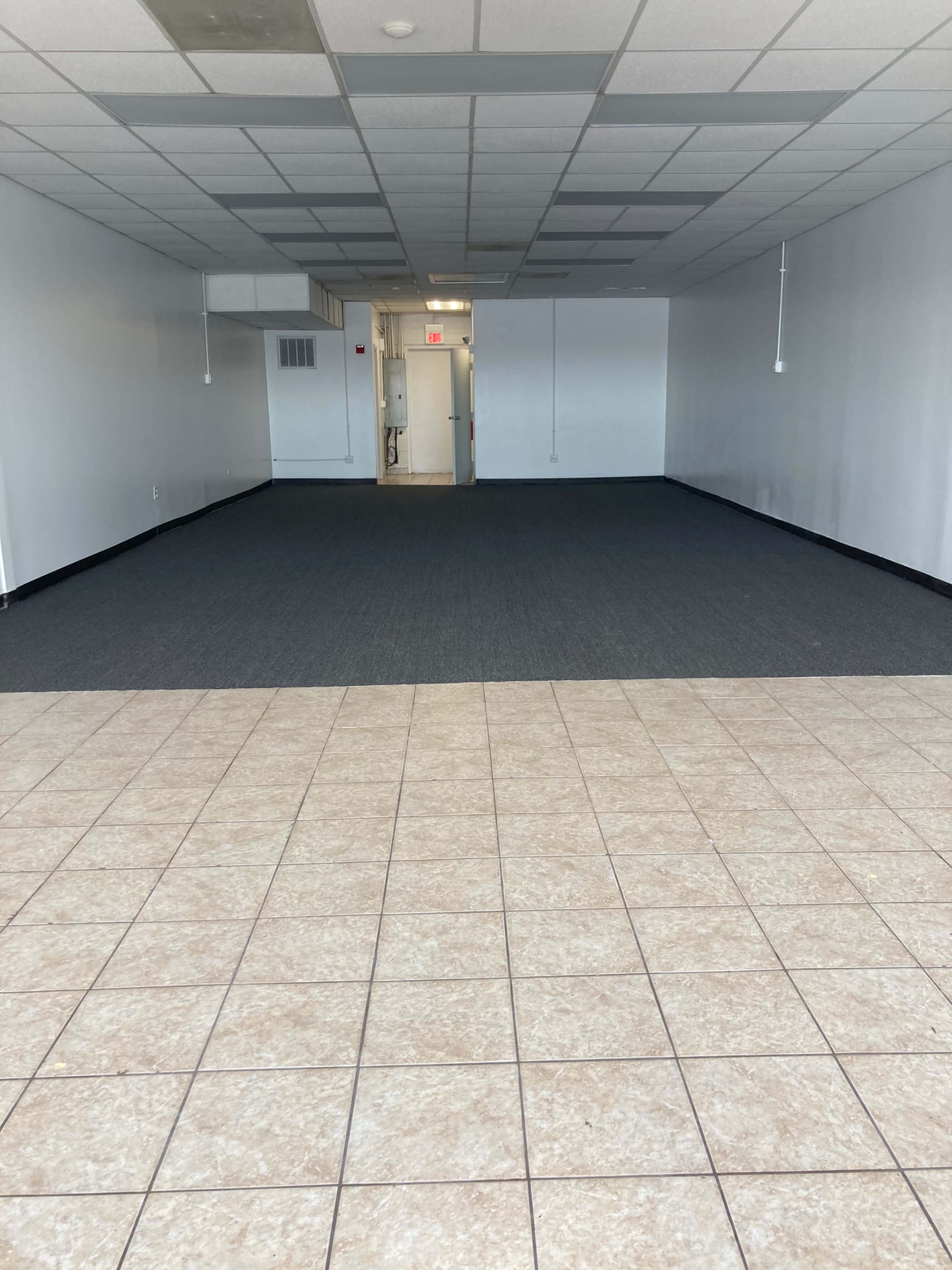 An empty room with tiled flooring in the foreground and carpeted flooring leading to a doorway at the back.