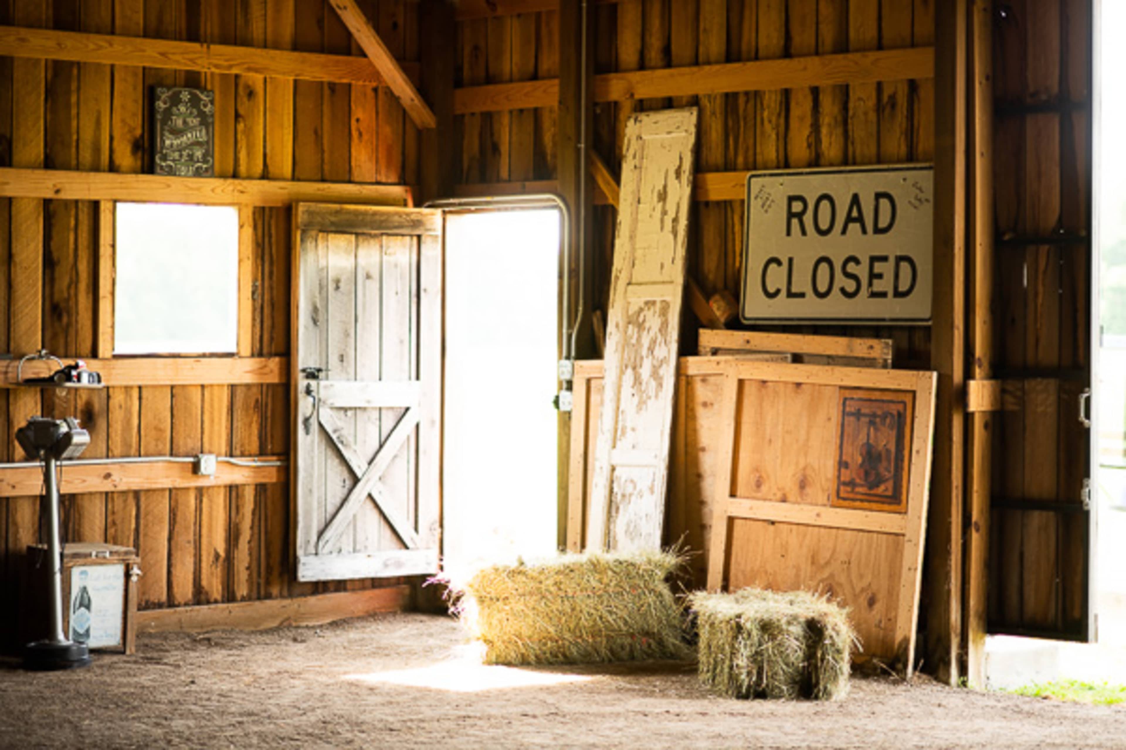 The interior of a wooden barn features hay bales, a closed door, and various signs propped against the walls.
