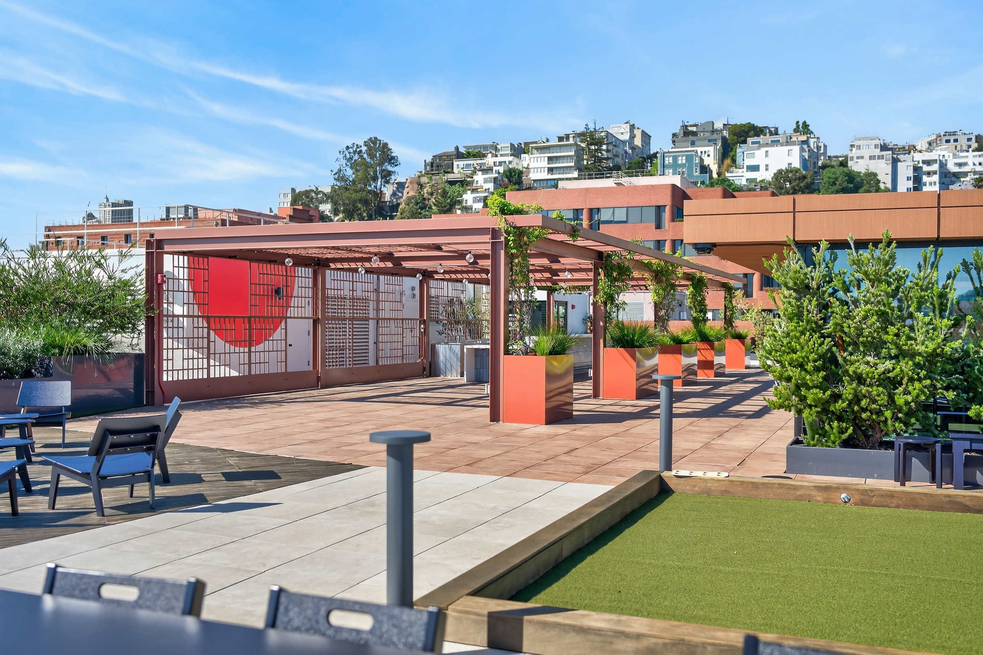 The image shows an outdoor rooftop space featuring a pergola structure, potted plants, and various seating arrangements with modern architecture in the background.