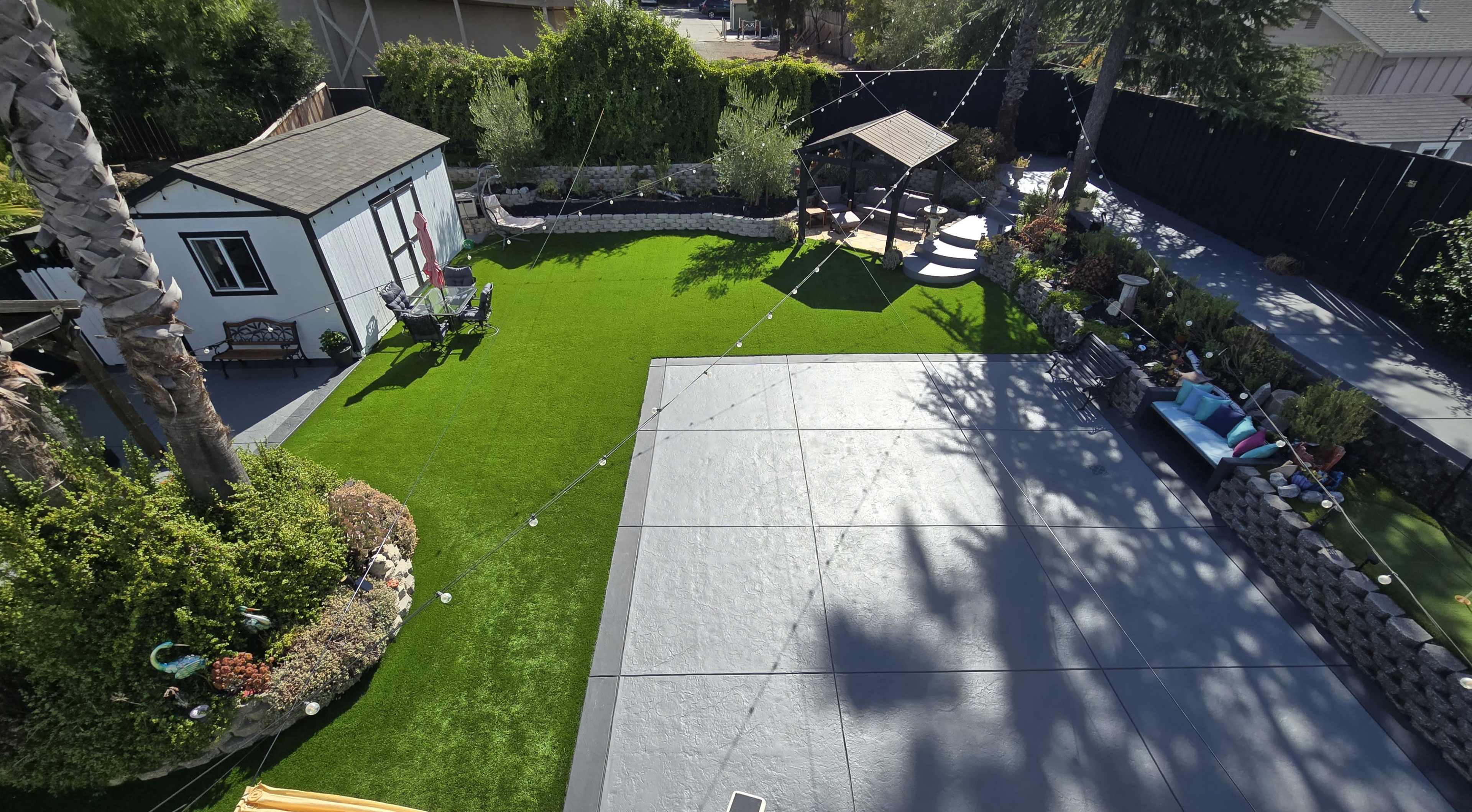 The image shows a landscaped backyard featuring a green synthetic lawn, a gazebo, a small shed, and various seating areas.