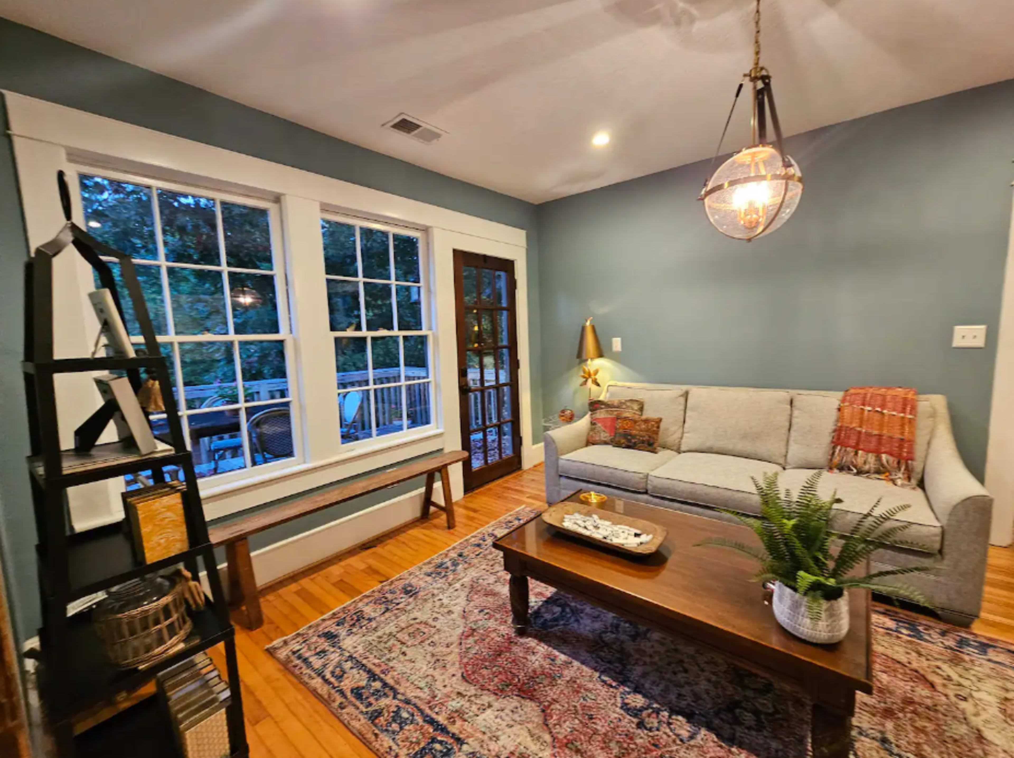 The scene features a cozy living room with a light gray sofa, a wooden coffee table, and large windows allowing natural light to illuminate the space.