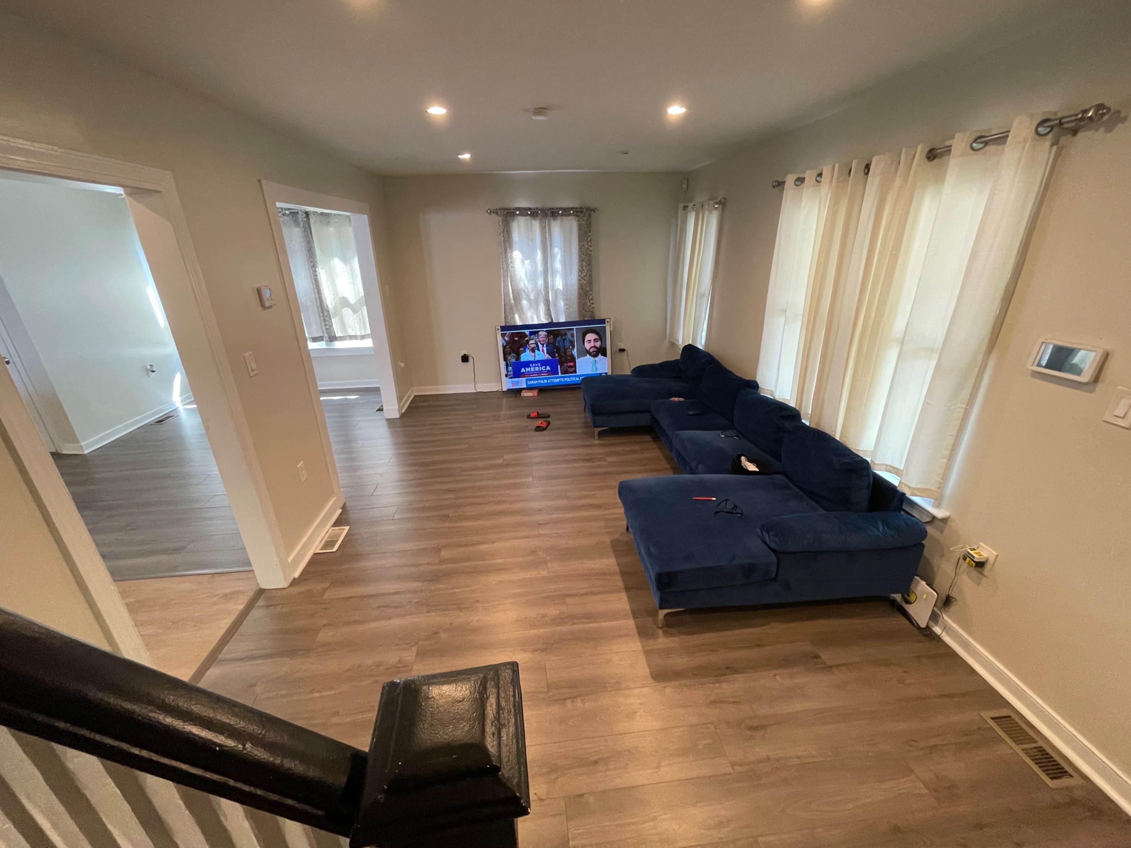 The image shows a living room with a blue sectional sofa facing a television on a wooden floor, with large windows covered by sheer curtains.
