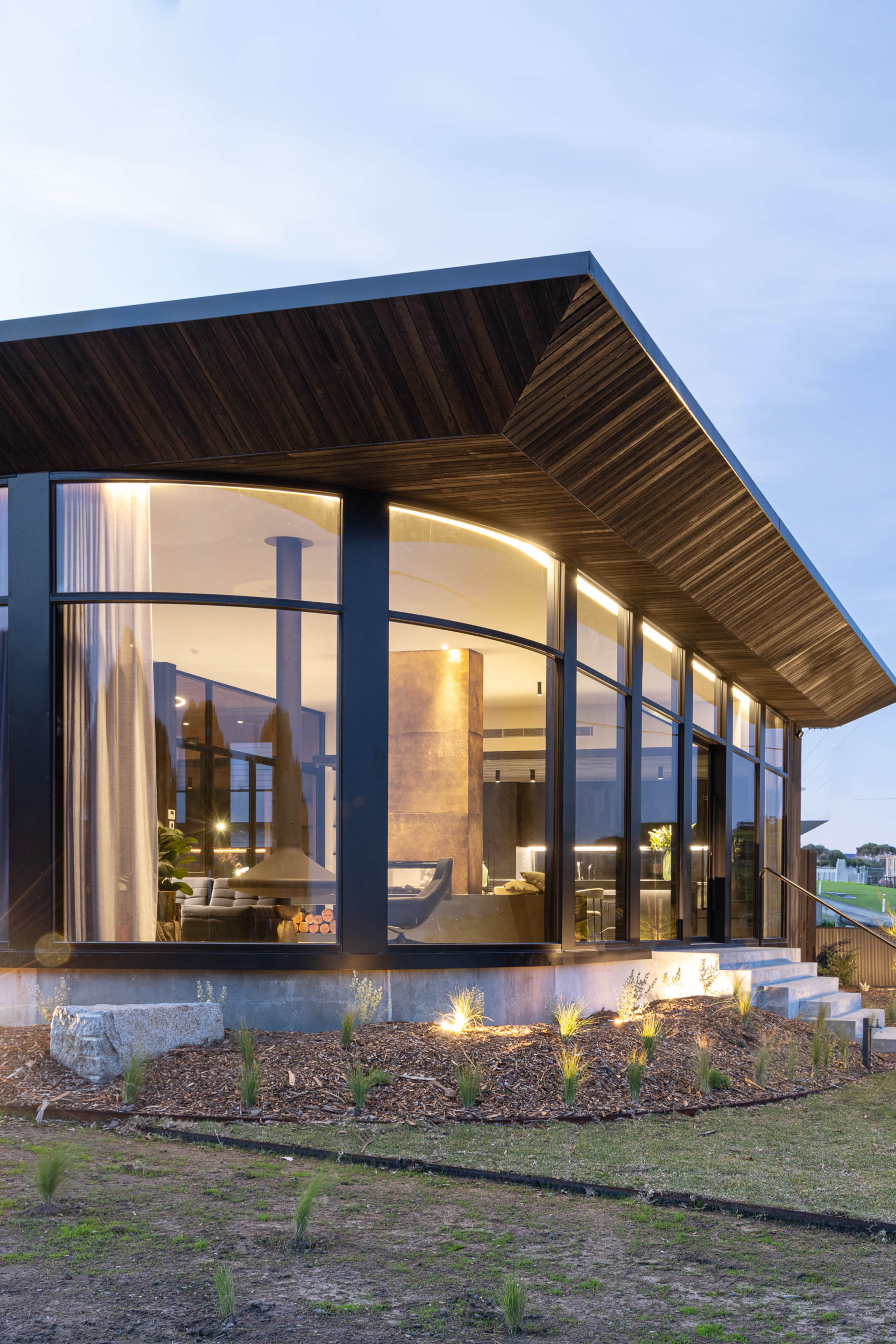 A modern house features a large angular roof and extensive glass windows, showcasing the interior and surrounding landscaping.