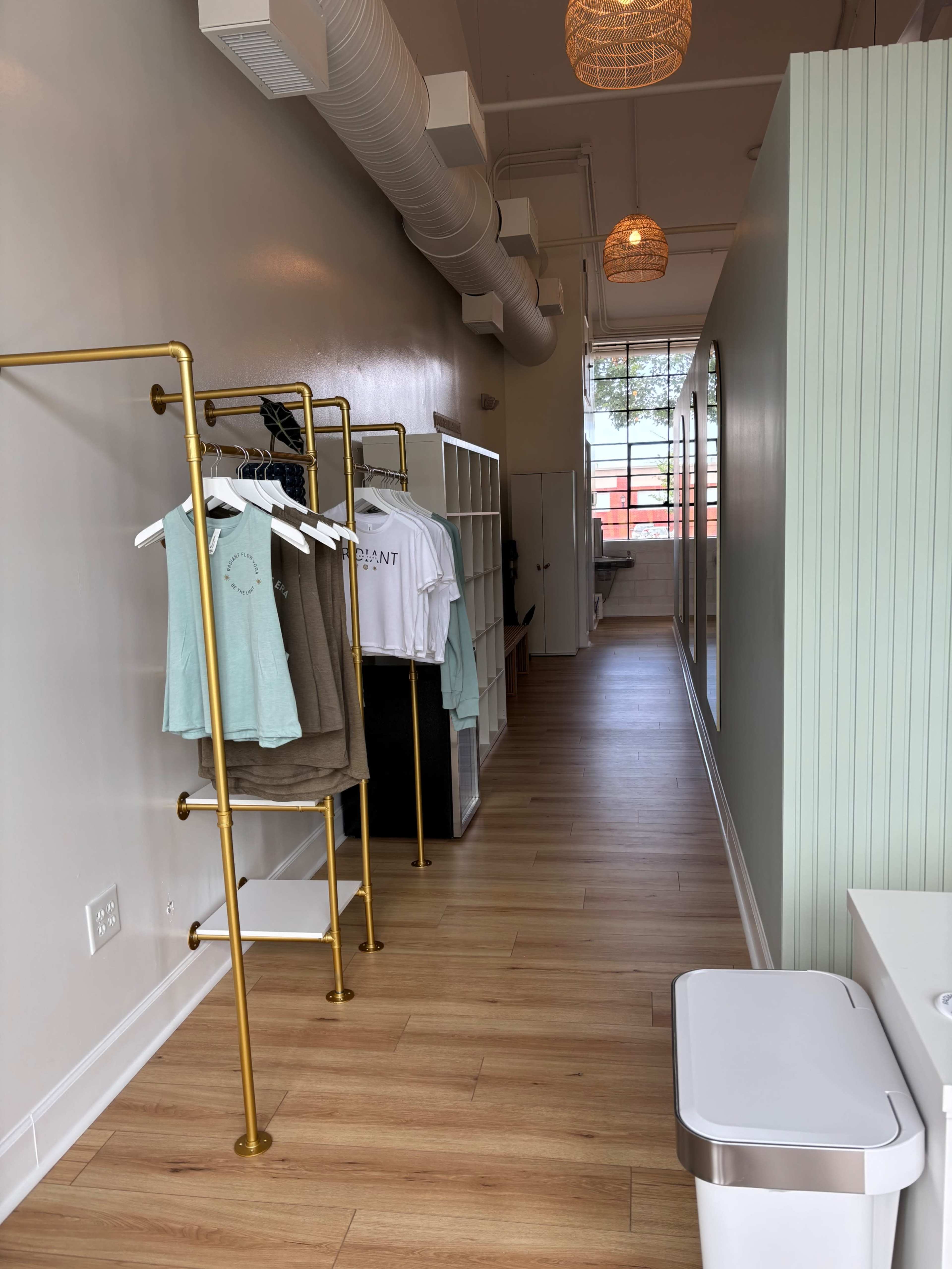 The image shows a narrow retail space with wooden flooring, light-colored walls, and clothing racks displaying garments along one side.