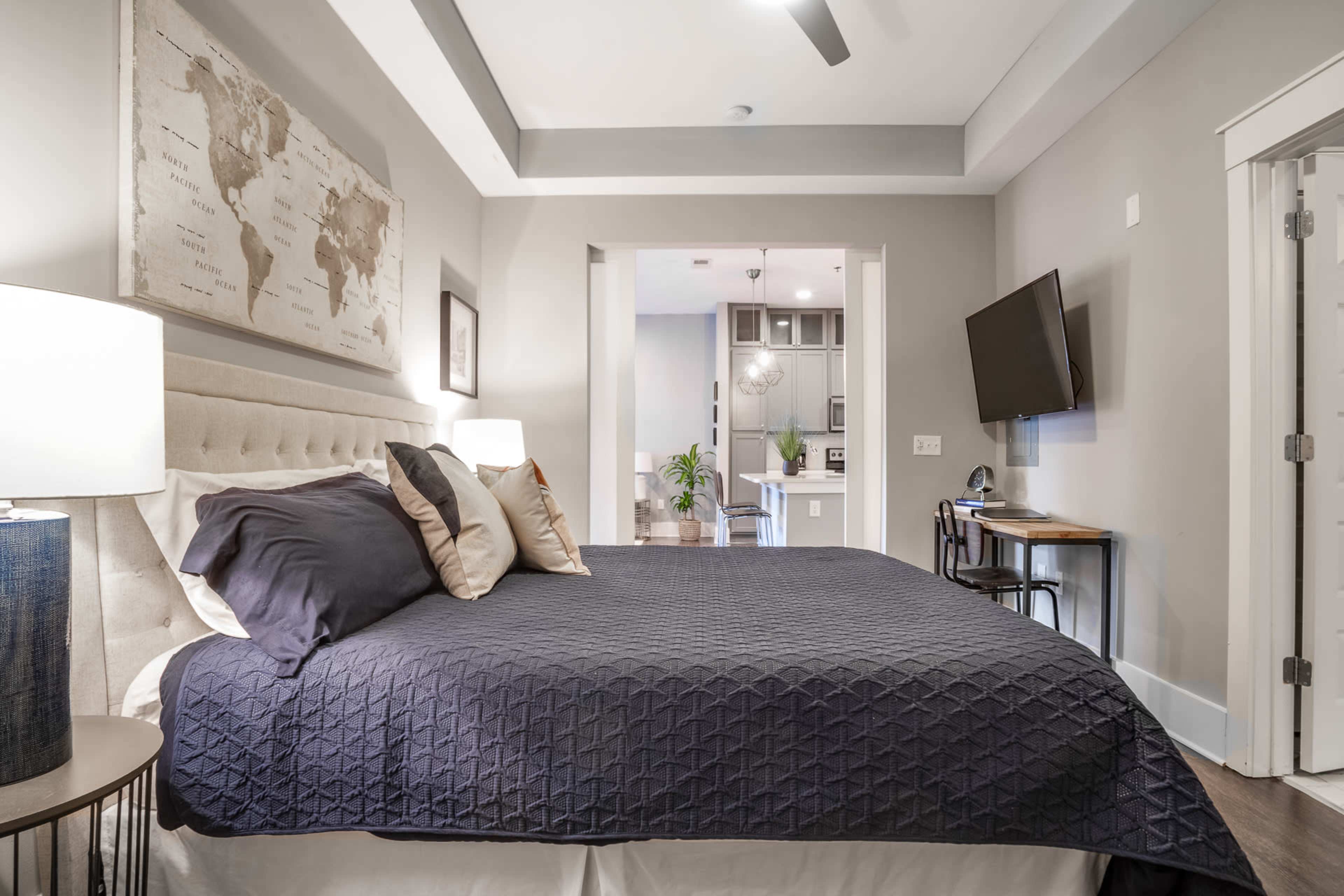 A well-furnished bedroom features a large bed with a dark quilt, a world map on the wall, and a small workspace with a TV and chair in the background.