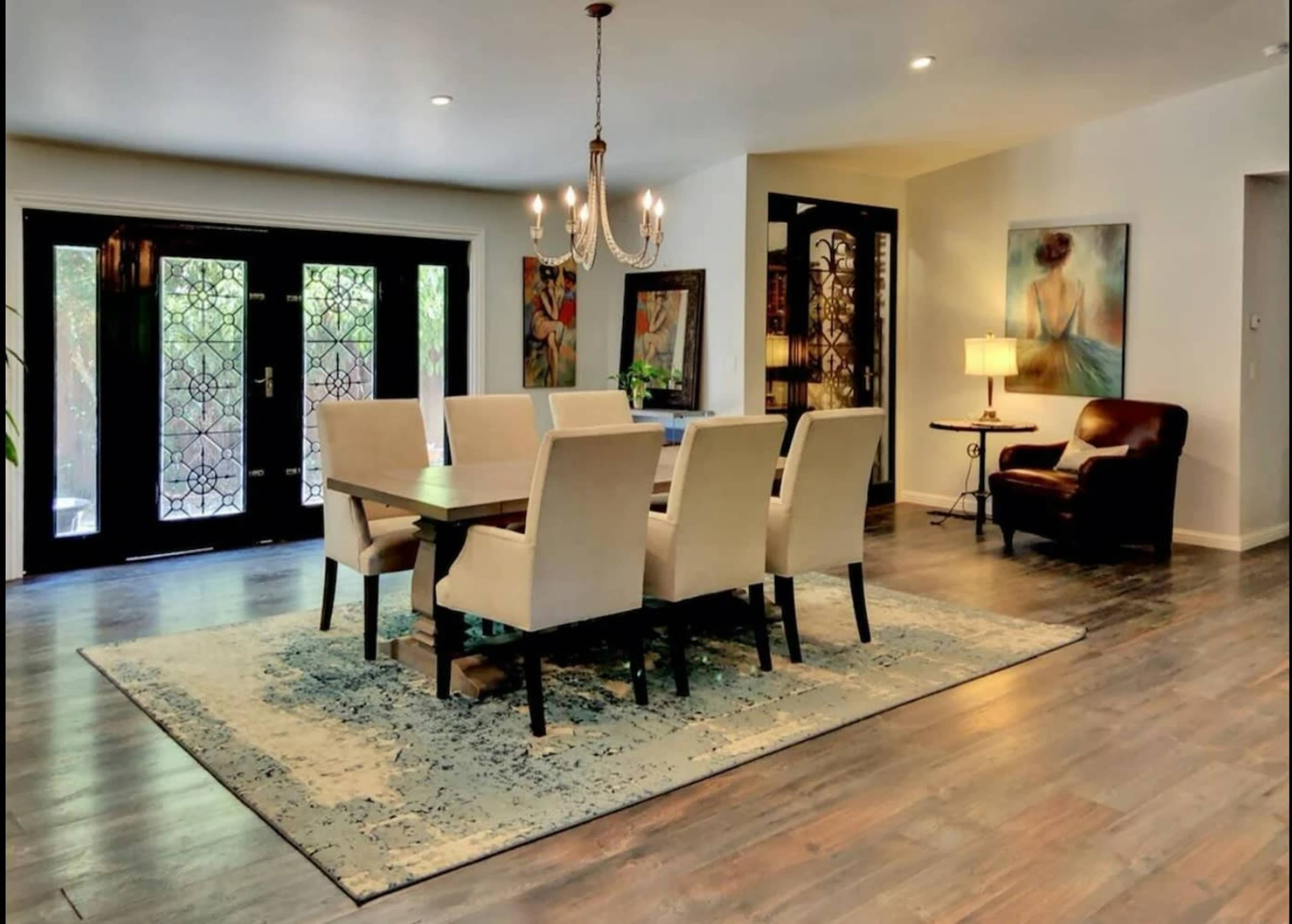 A dining area with a large wooden table surrounded by beige chairs, a chandelier overhead, and glass doors leading to an outdoor space.