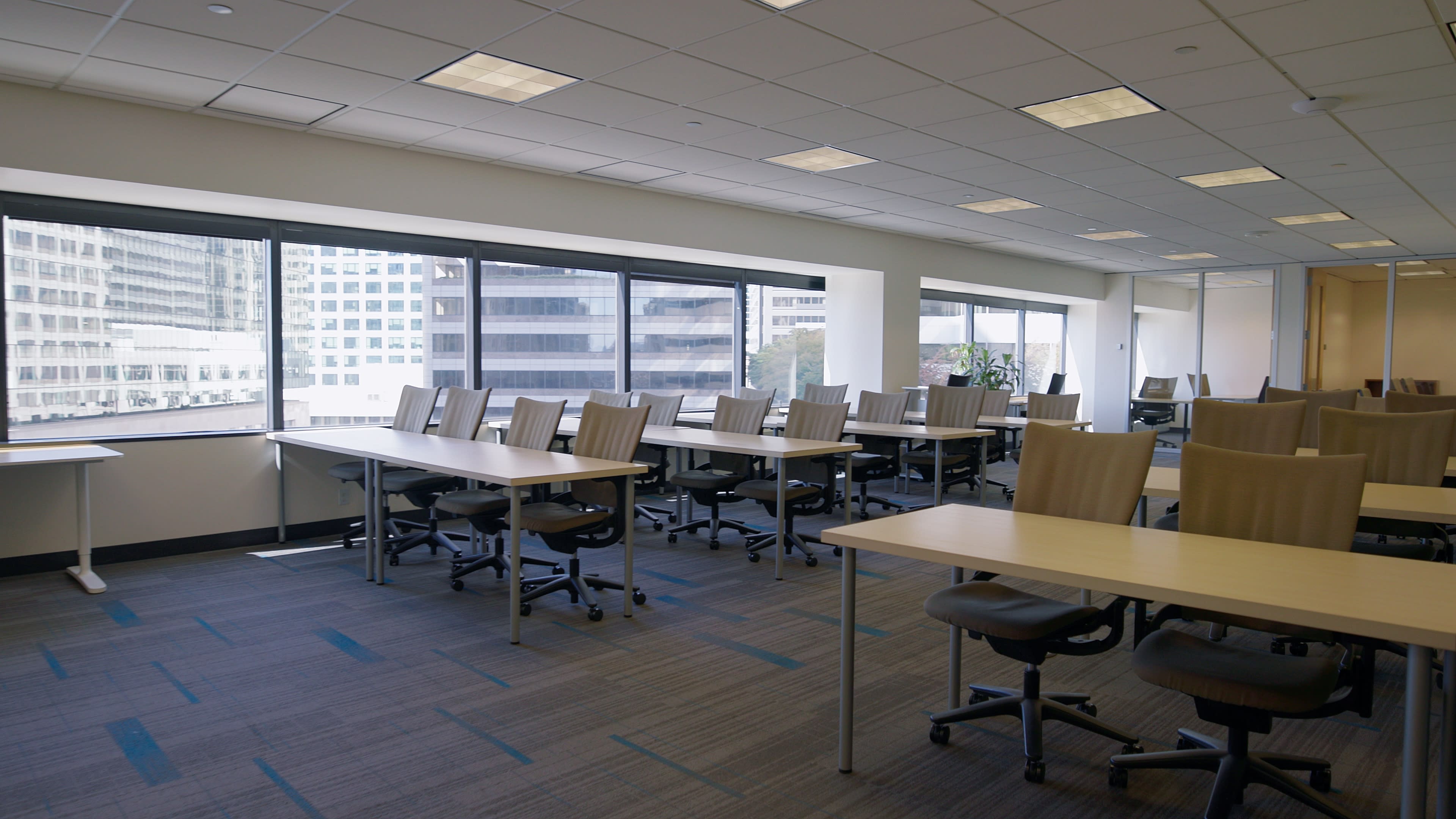 The image shows a modern office space with rows of empty desks and large windows providing views of a cityscape.