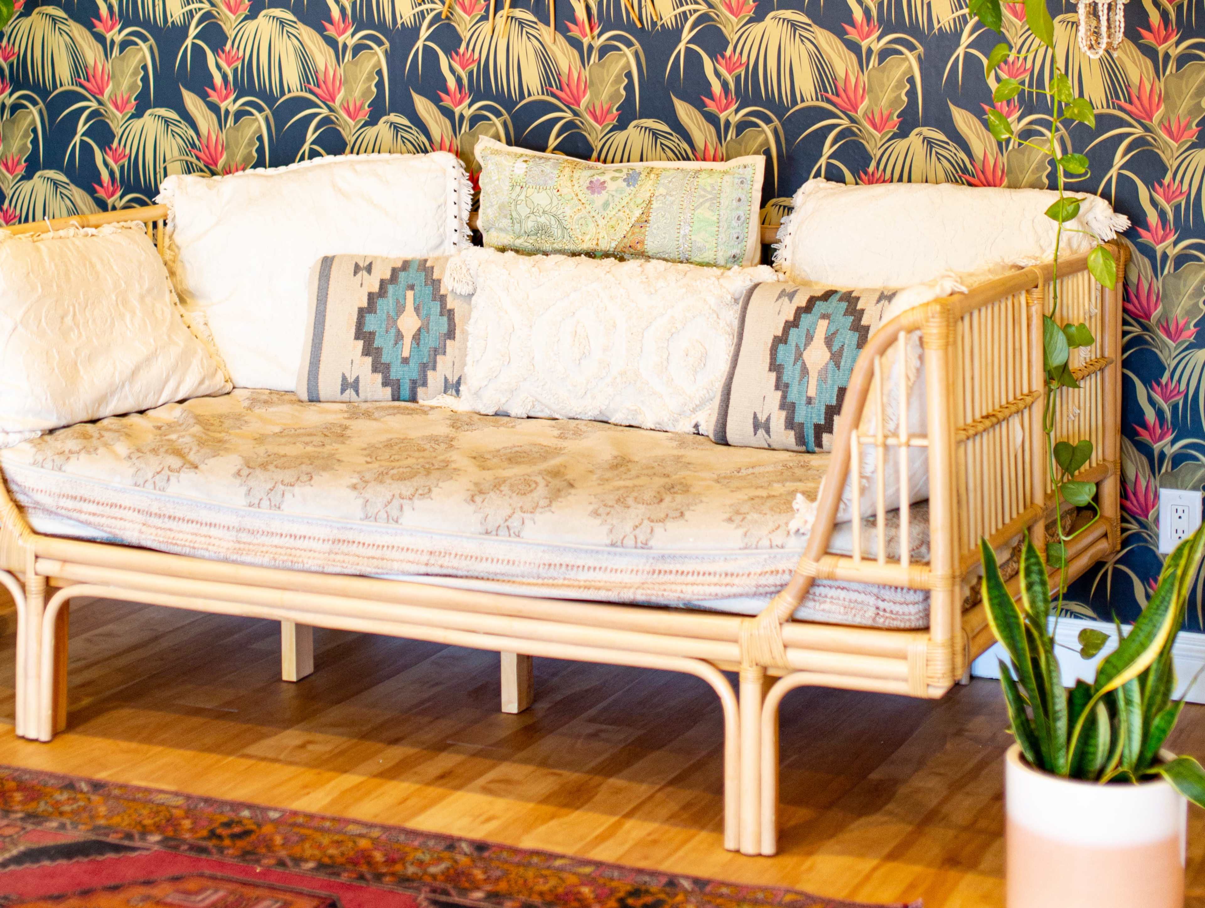 A rattan sofa with decorative pillows is placed against a floral-patterned wallpaper in a well-lit room.
