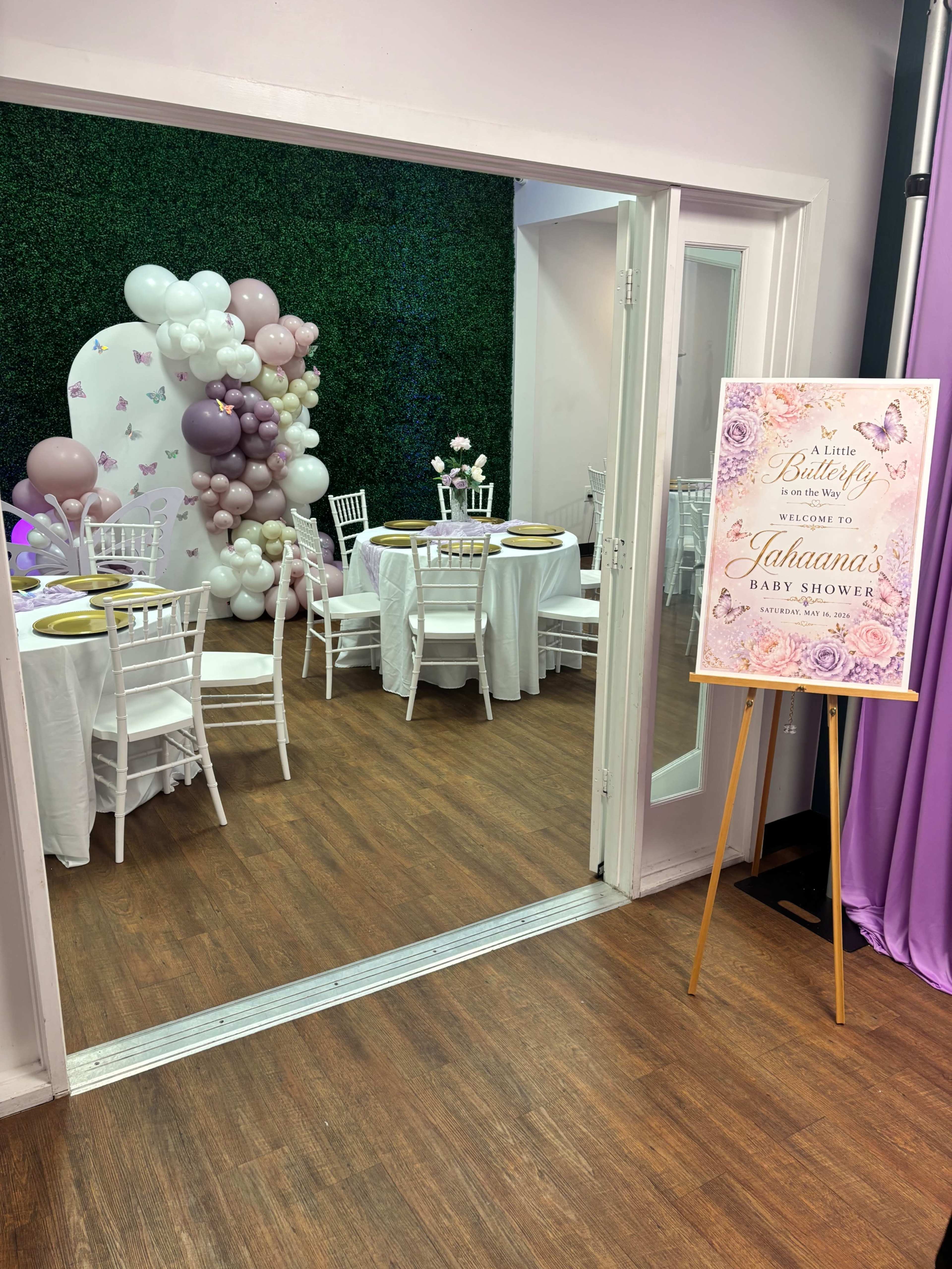 A decorated room for a baby shower features a green hedge backdrop, balloons, and tables set with white chairs.