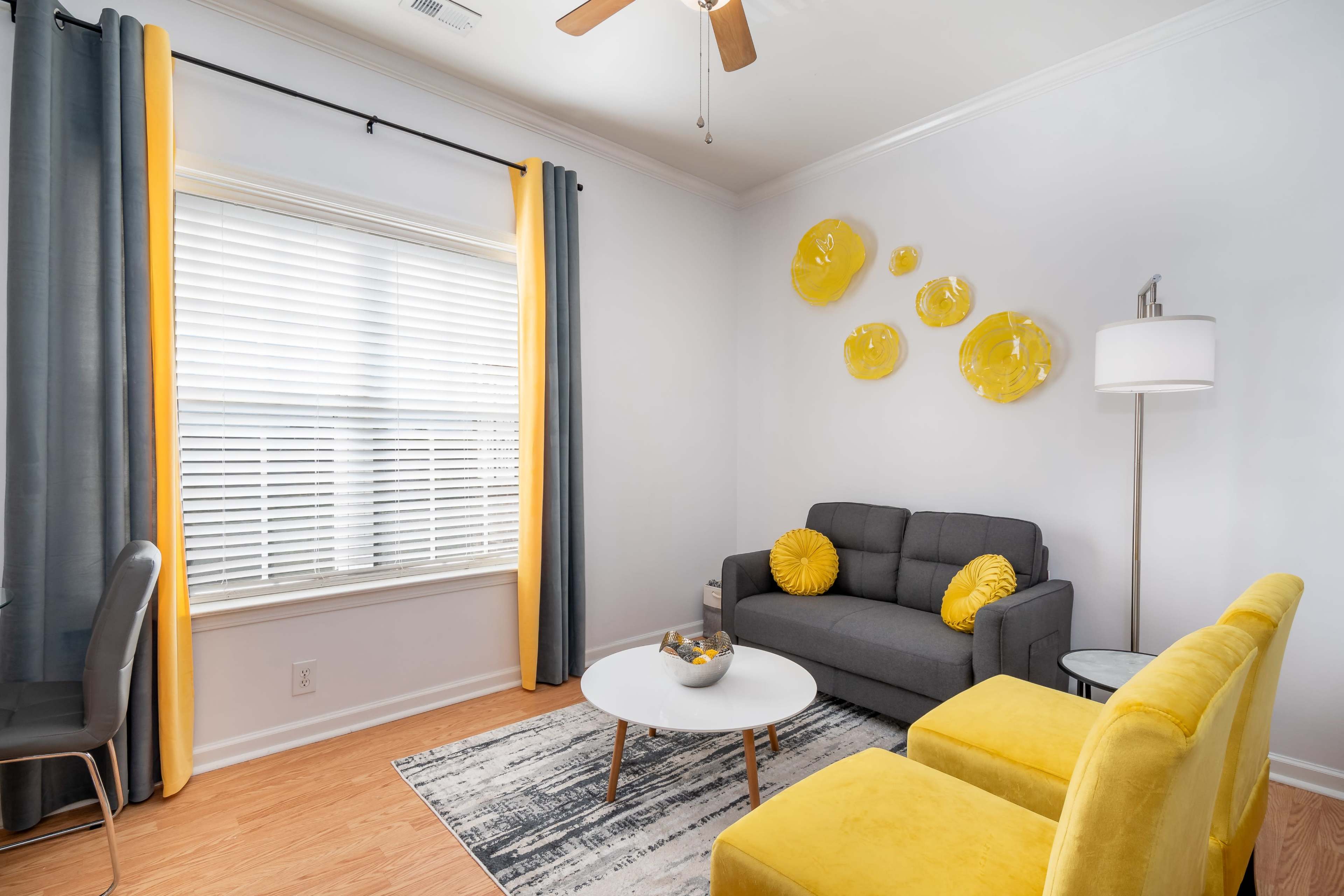 A living room with a gray couch and two yellow armchairs, a round coffee table, and yellow wall decor, all complemented by bright window light.