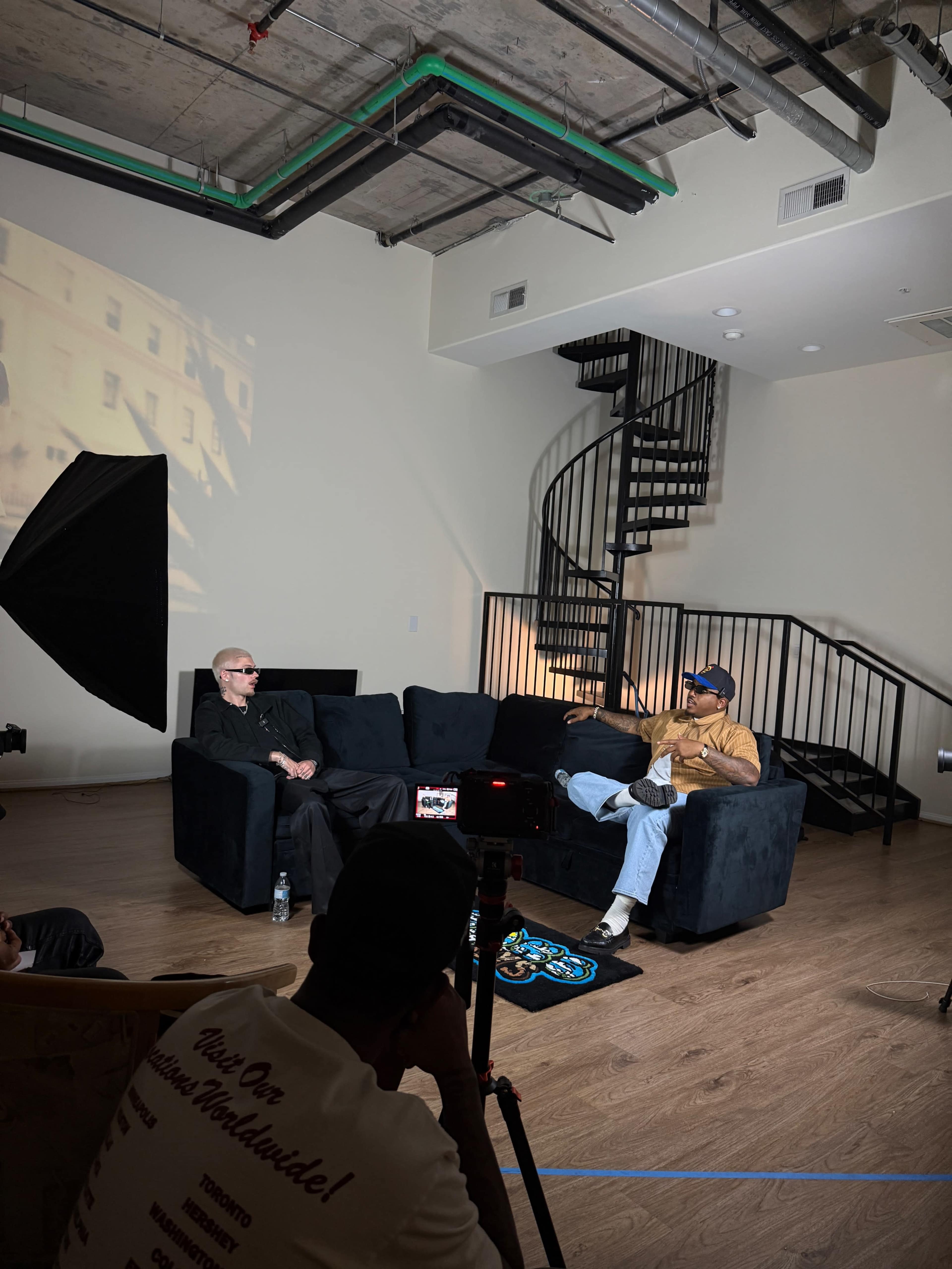 Two men are seated on a black couch in a modern room with a spiral staircase, while a camera and lighting equipment are set up for a shoot.