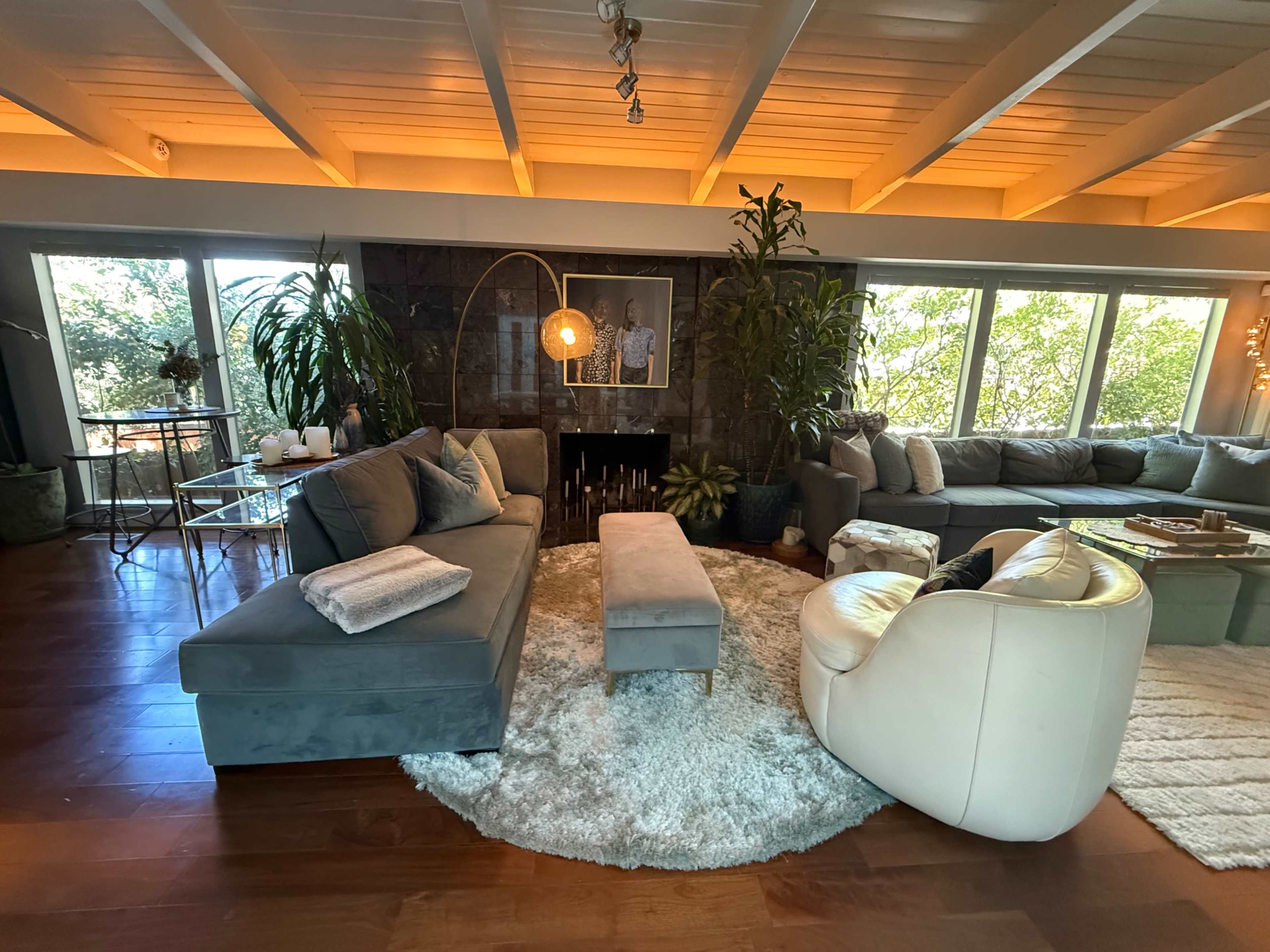 The living room features a sectional sofa, a curved armchair, a coffee table, and large windows with greenery visible outside.