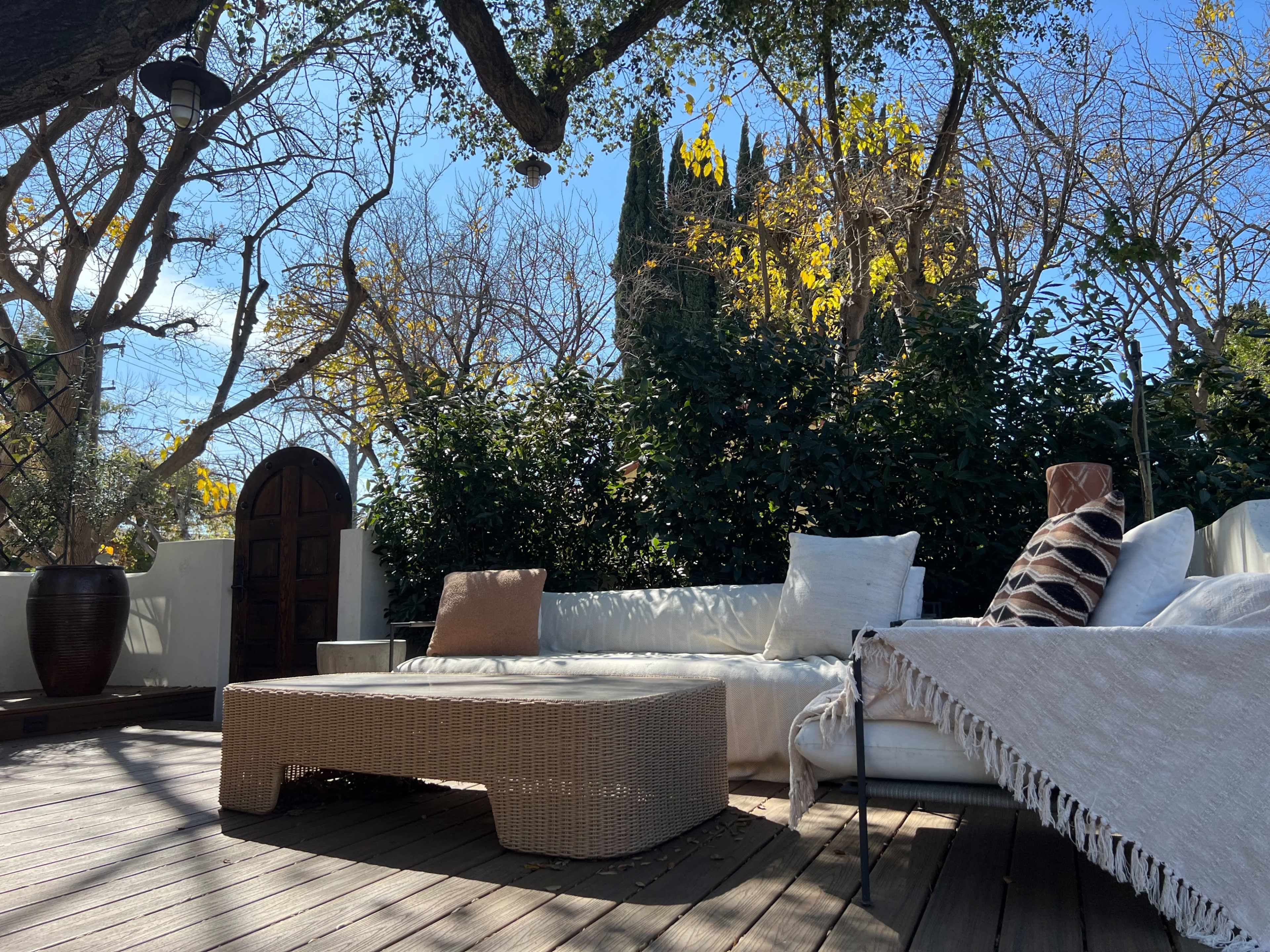 A spacious outdoor seating area features a wicker sofa and a wooden deck surrounded by trees and greenery.