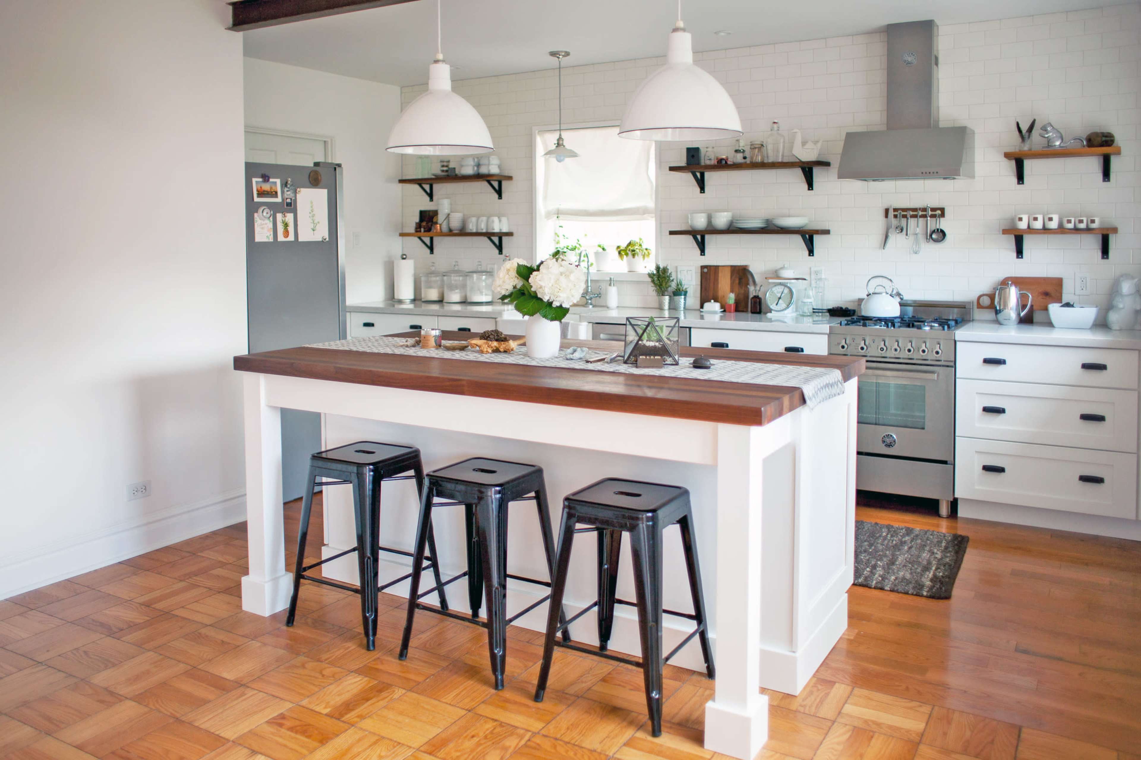 A modern kitchen with white cabinetry, a wooden island, and metal bar stools, featuring open shelves displaying dishware and a stove with a hood above.