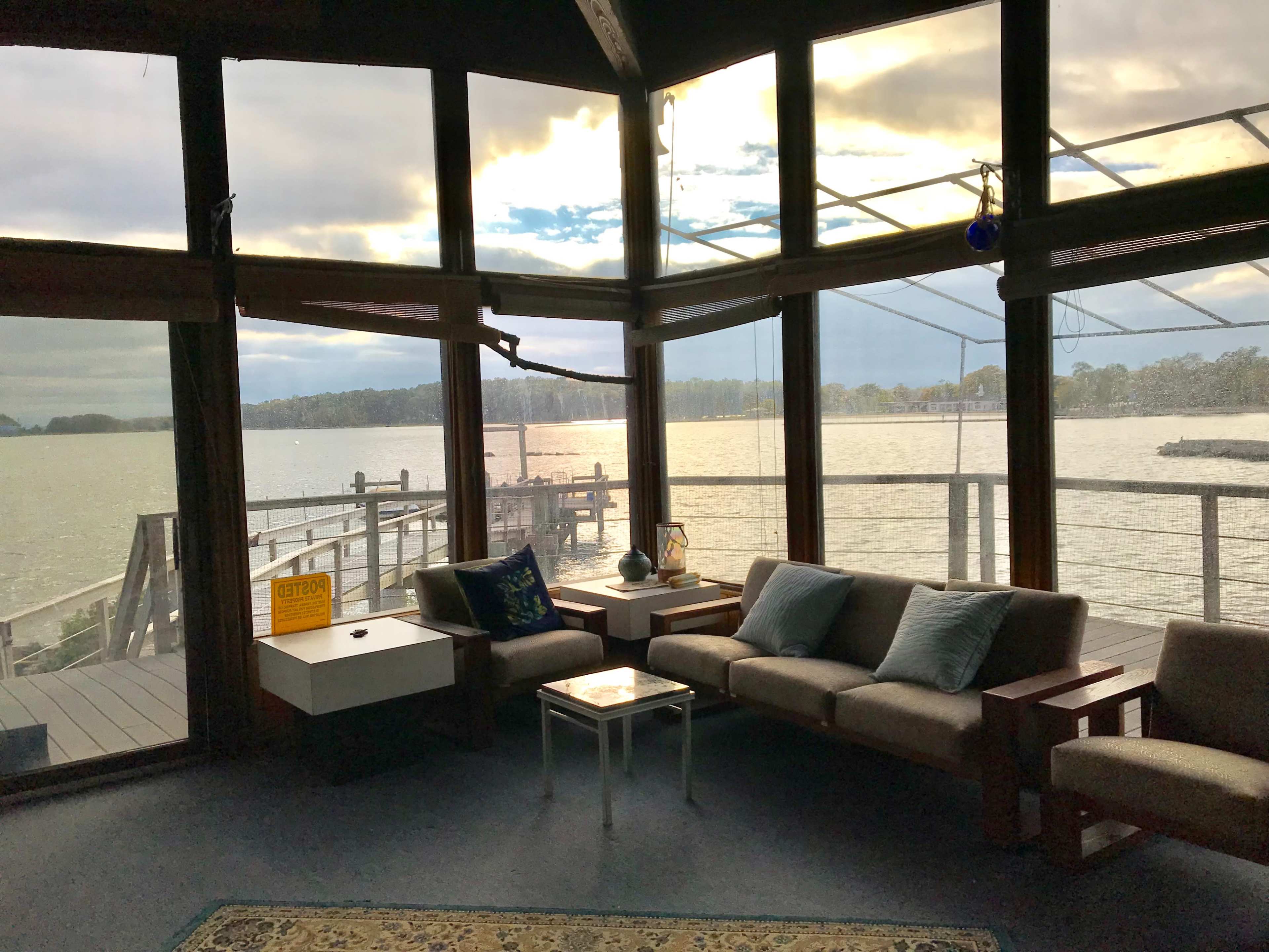 A spacious room with large windows offers a view of a lake and a dock, featuring modern furniture and a small table in the foreground.