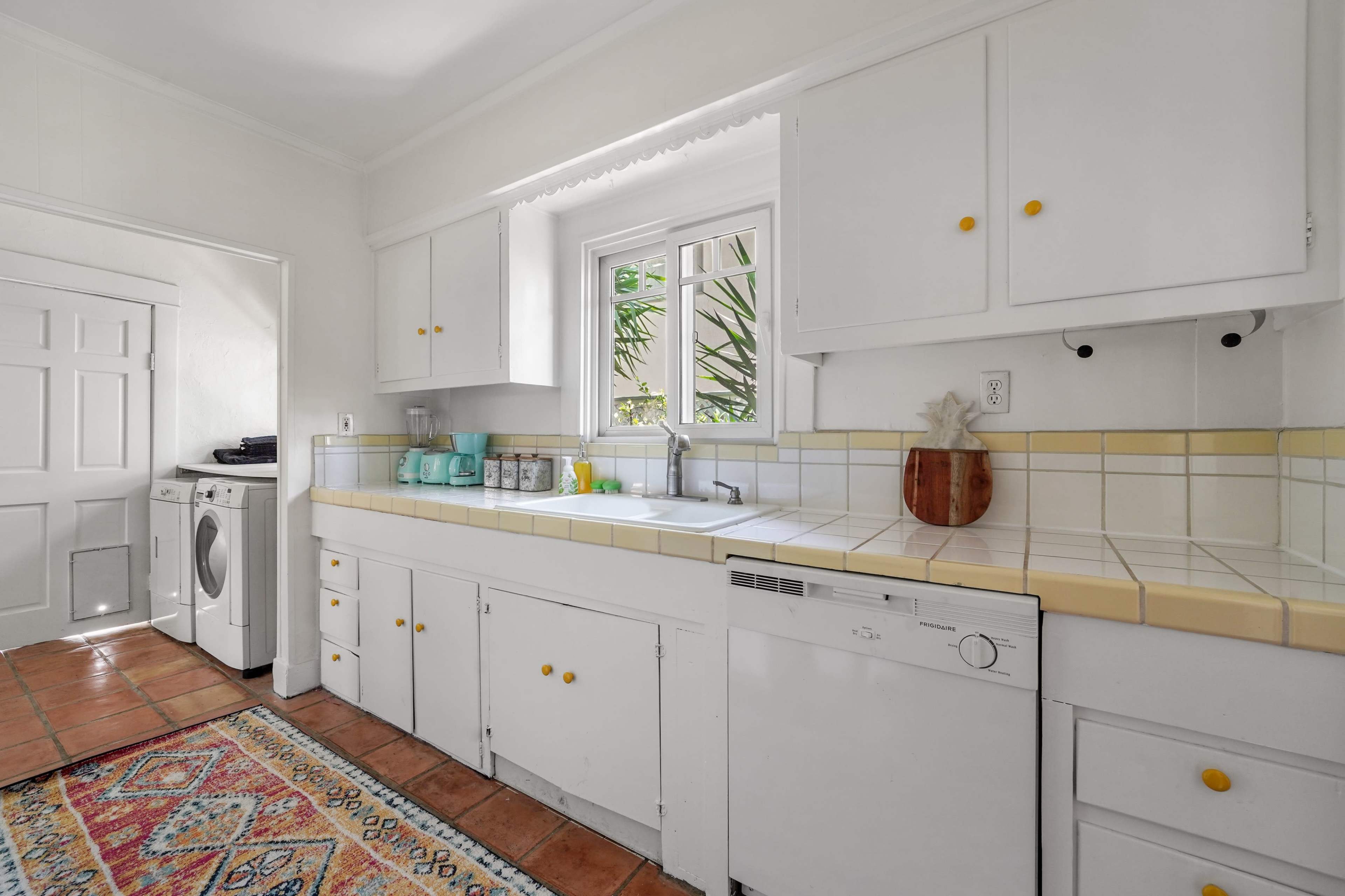 A bright kitchen features white cabinets, yellow knobs, and a ceramic sink beneath a window, with a washer-dryer unit and colorful rug visible in the space.