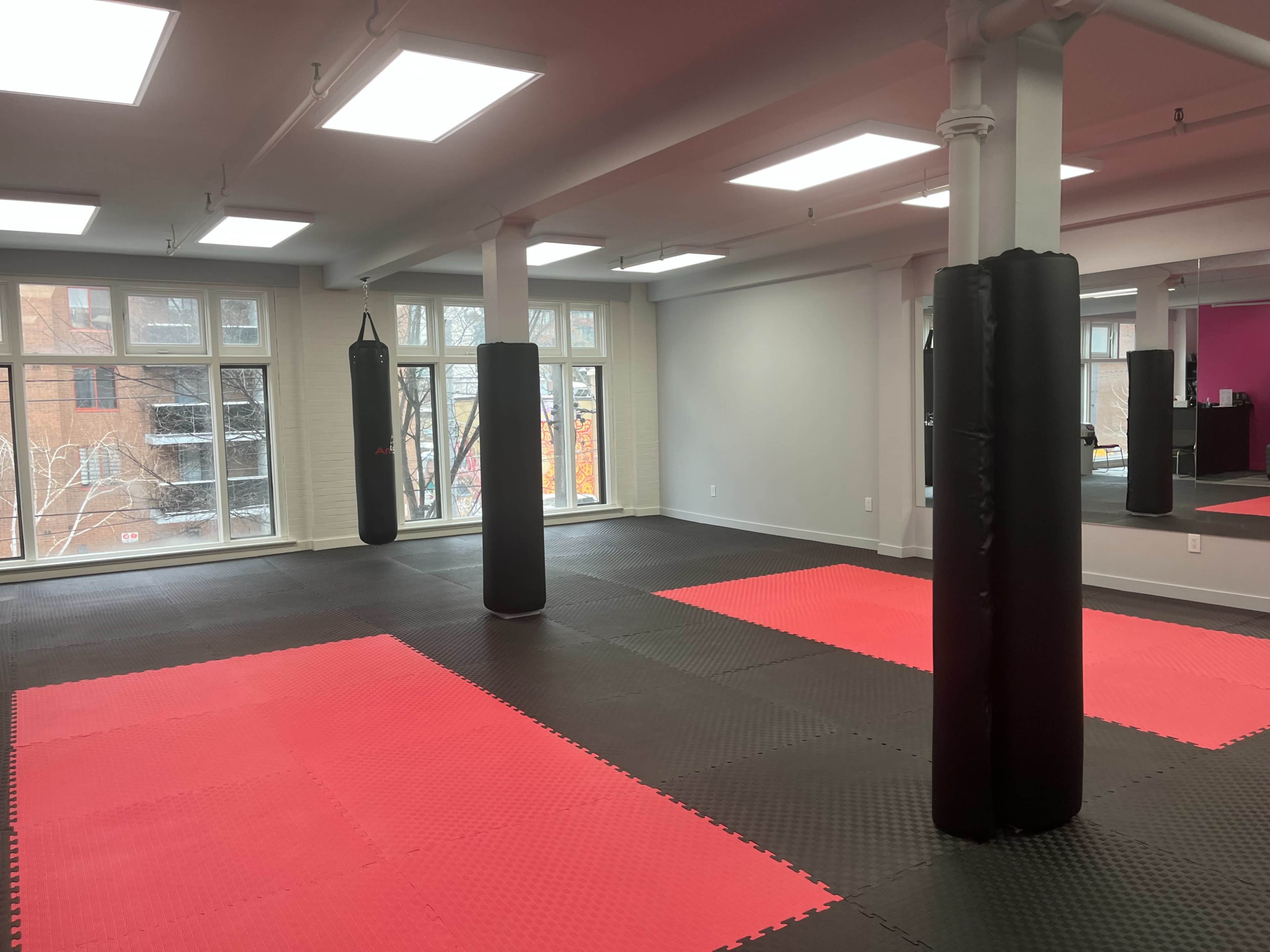 The image shows a spacious martial arts training studio with black punching bags and red mat flooring, illuminated by large windows.