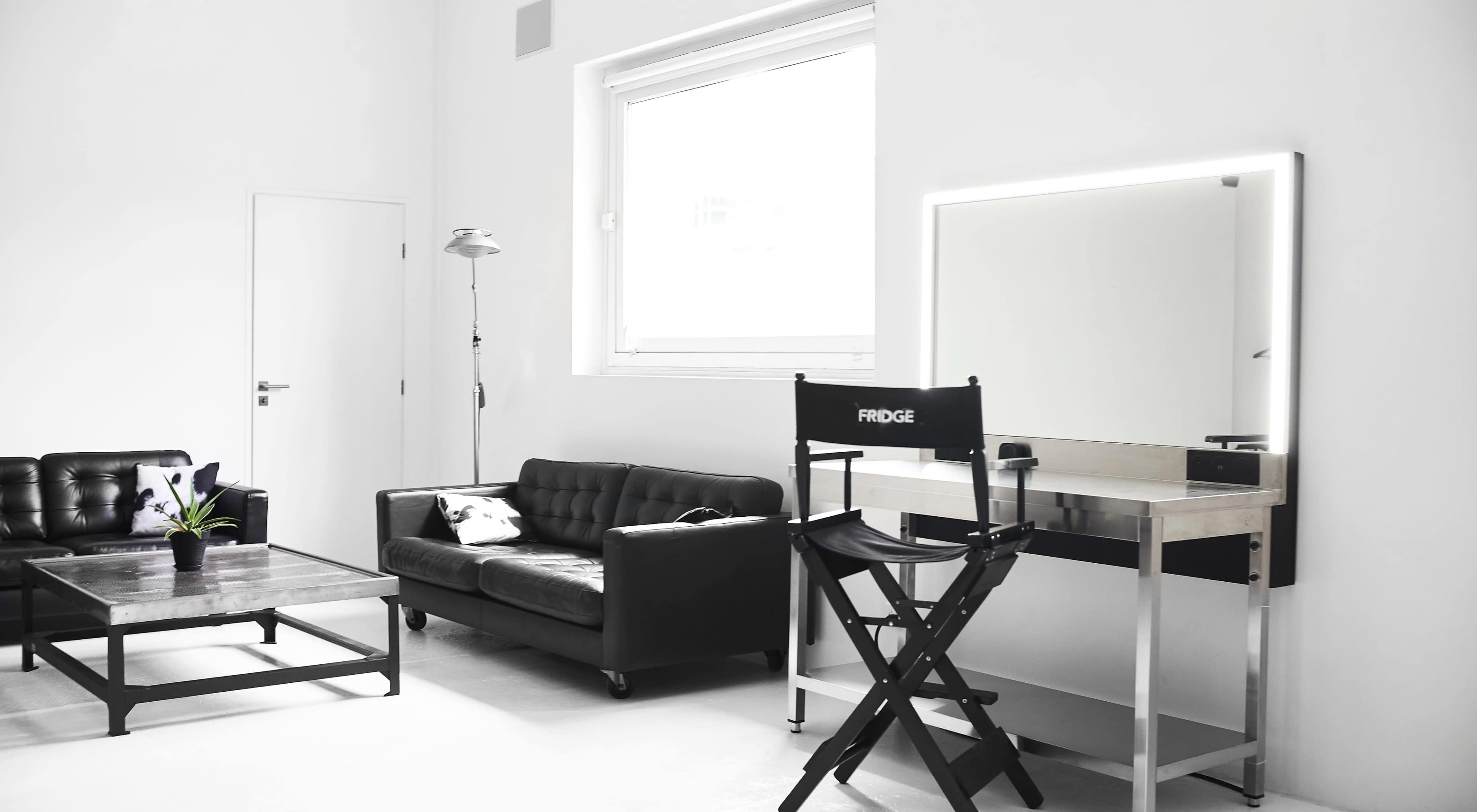 A modern, minimalist room featuring a black leather couch, a coffee table, a makeup vanity with a mirror, and a director's chair labeled "FRIDGE."