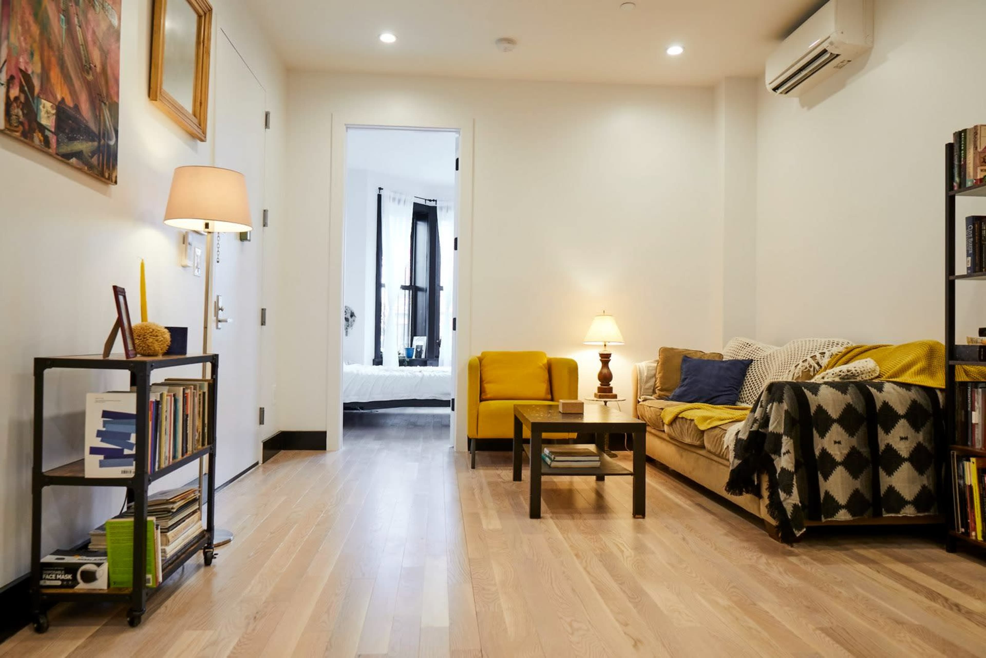 A living room with a yellow sofa, a wooden coffee table, a bookshelf, and an open door leading to a bedroom.