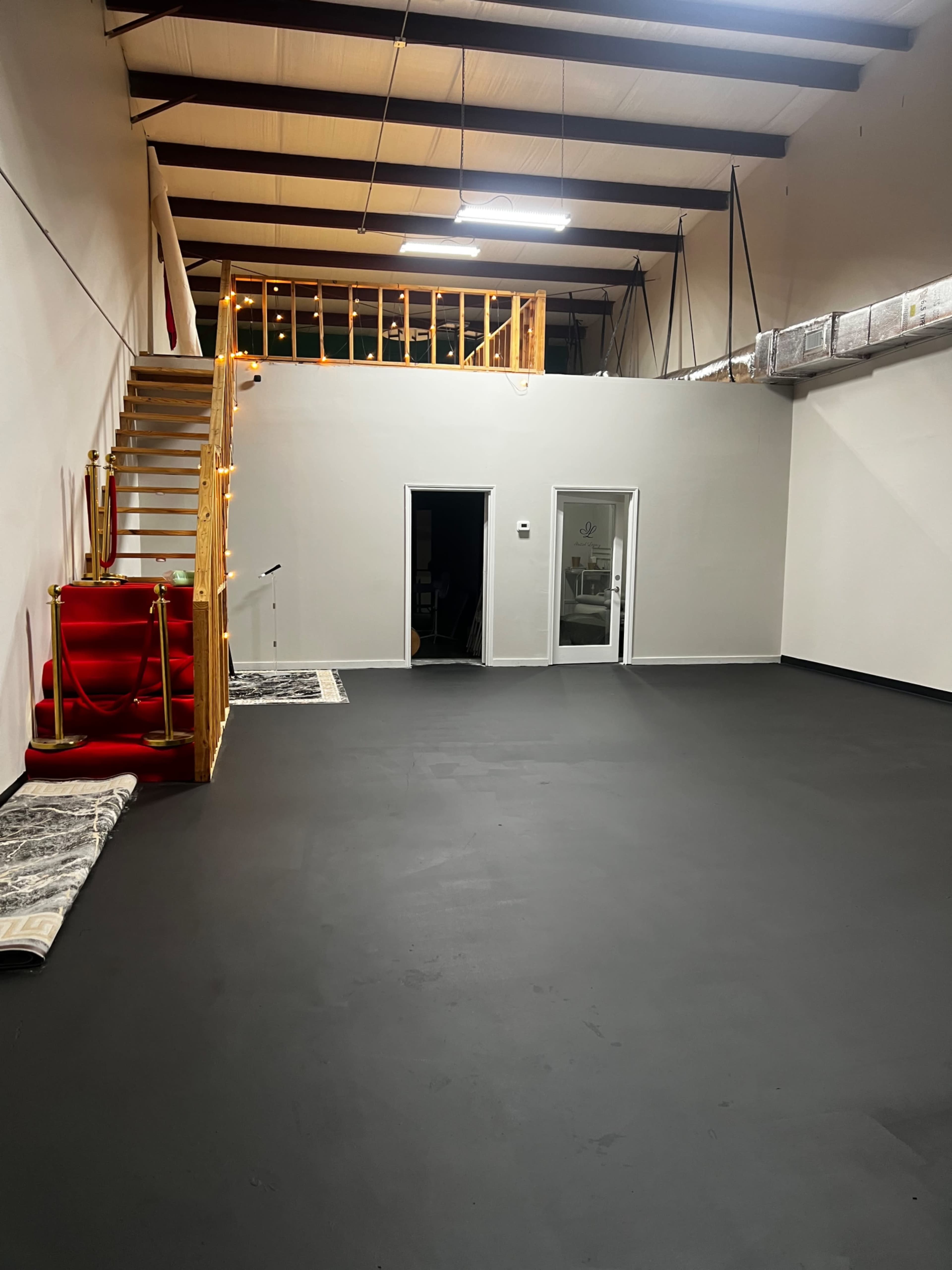 The image shows a spacious, empty room with a black floor, a staircase leading to a second level, and two doorways on one wall.