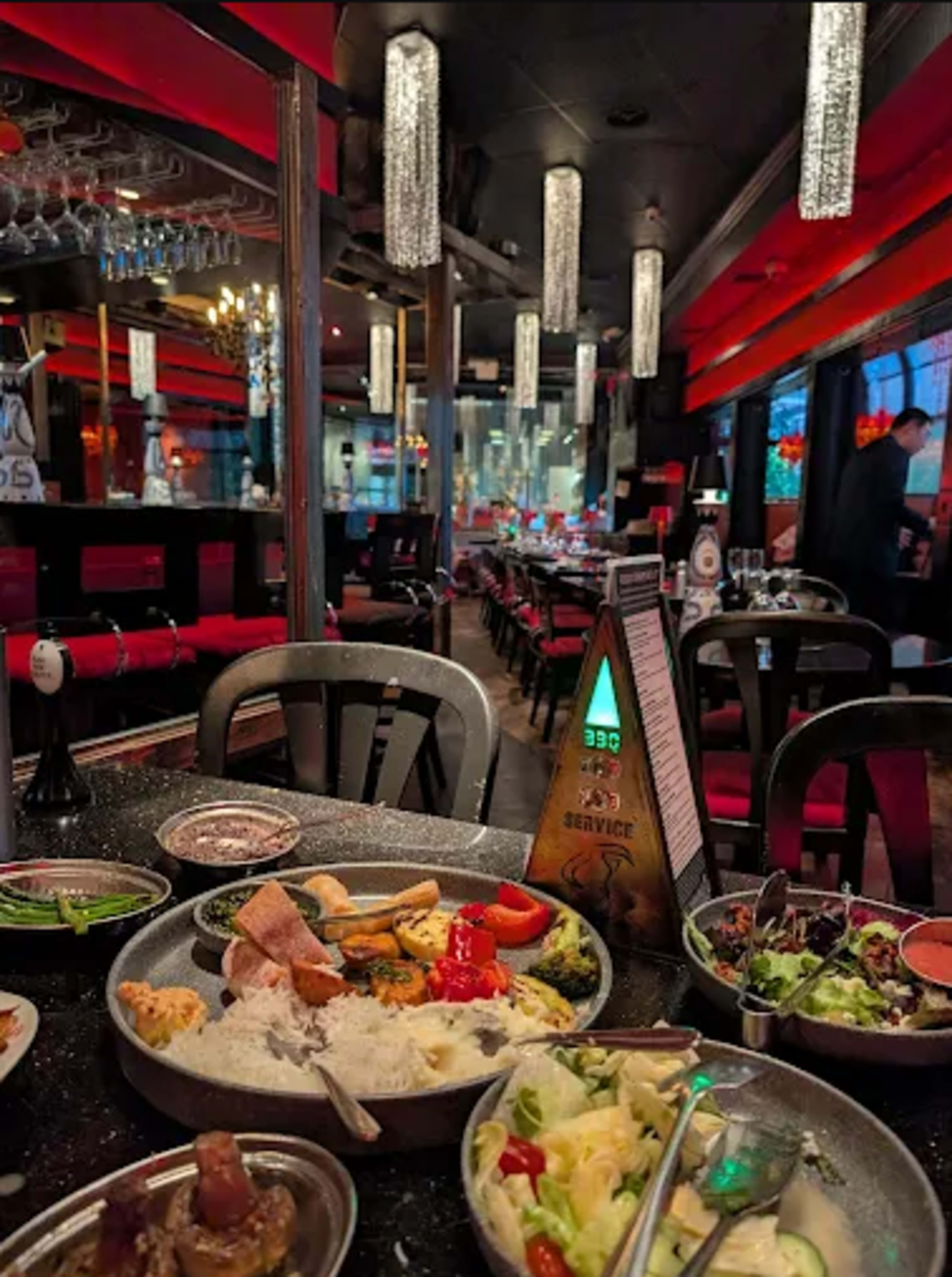 The image shows a table in a restaurant with multiple dishes of food, surrounded by a dimly lit interior featuring red accents and hanging light fixtures.