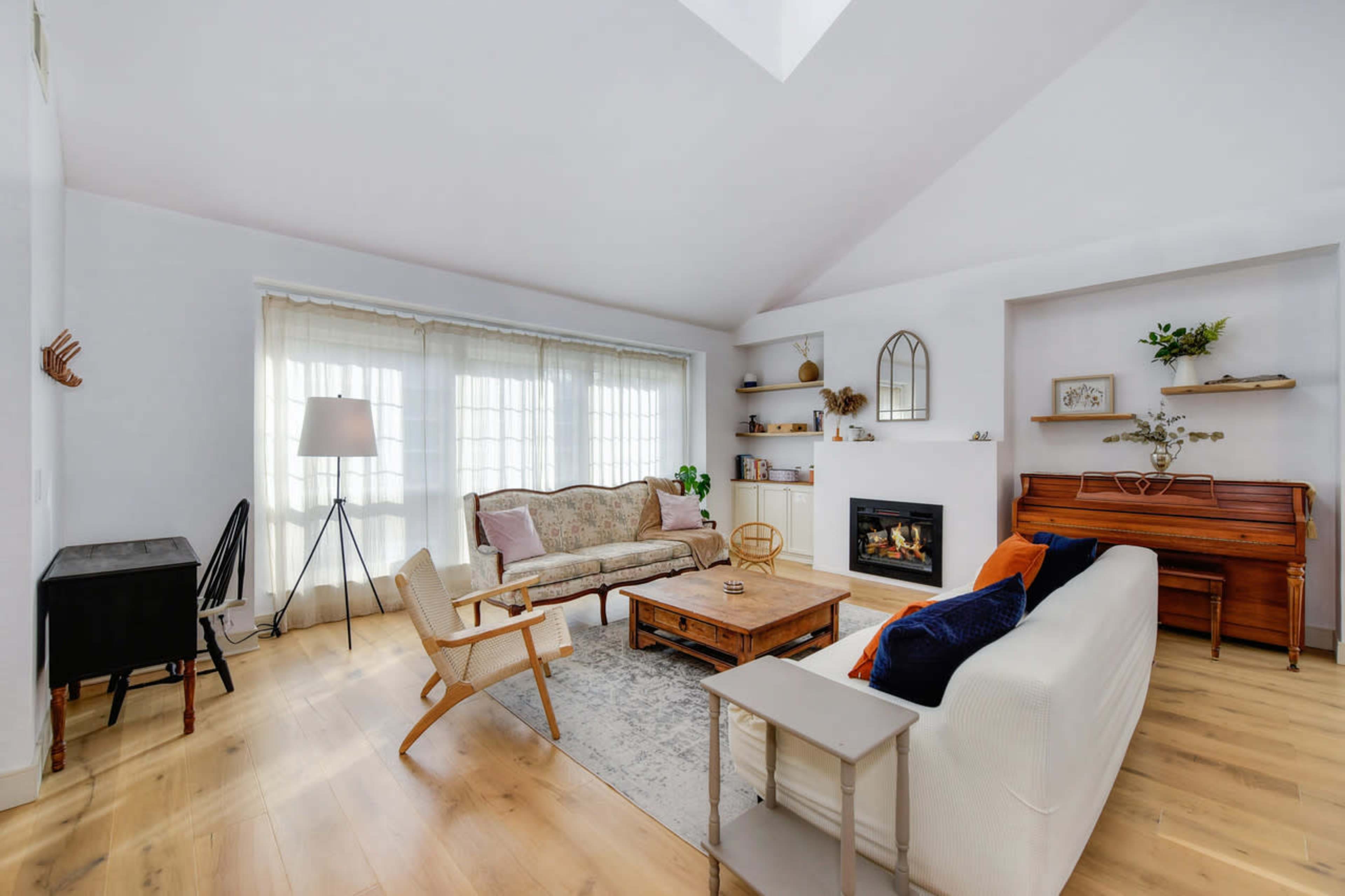 A spacious living room features a mix of modern and vintage furniture, including a sofa, armchairs, a coffee table, and a piano, with large windows allowing natural light to fill the space.