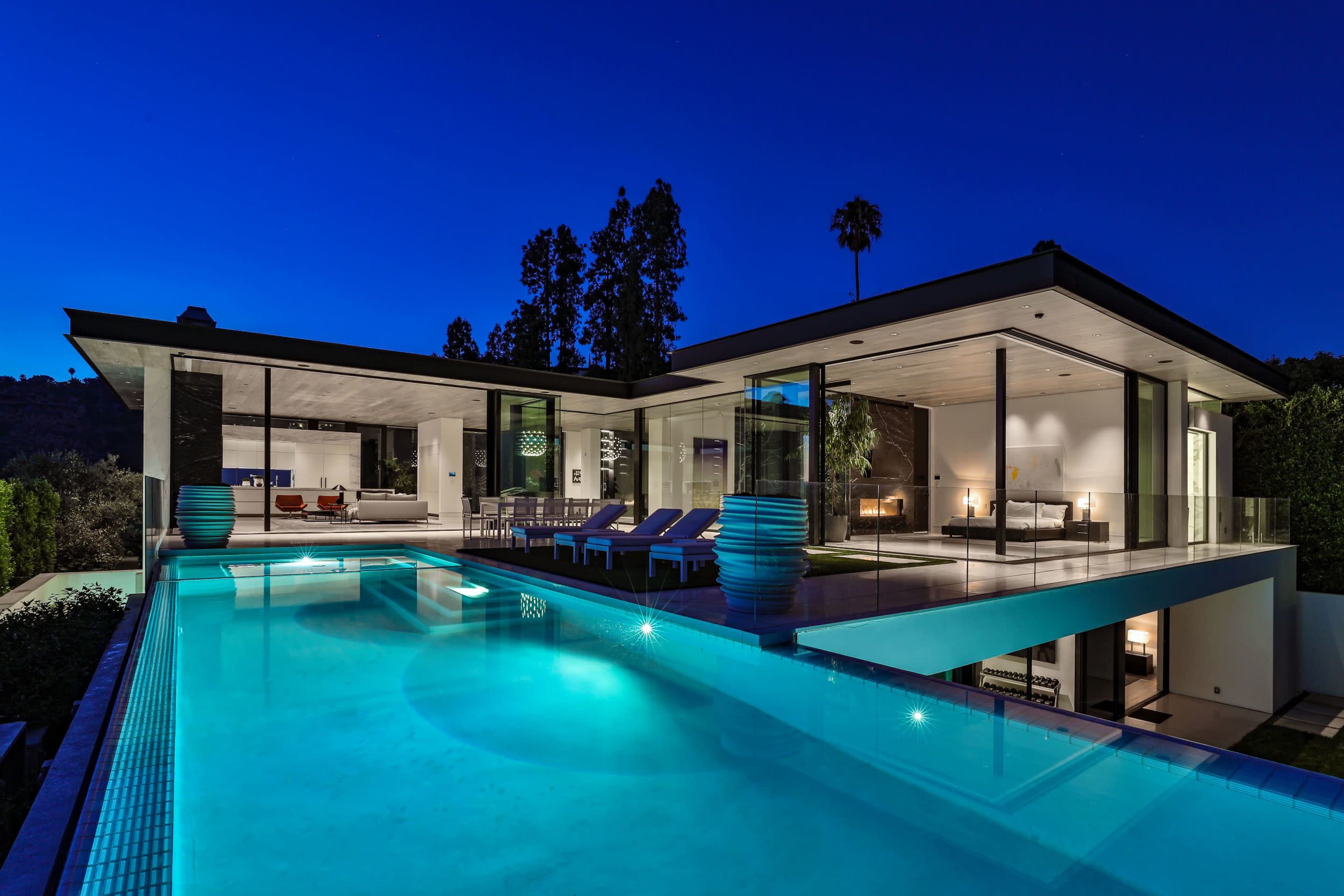 A modern home features a sleek pool area illuminated at night, with large glass walls revealing the luxurious interior.