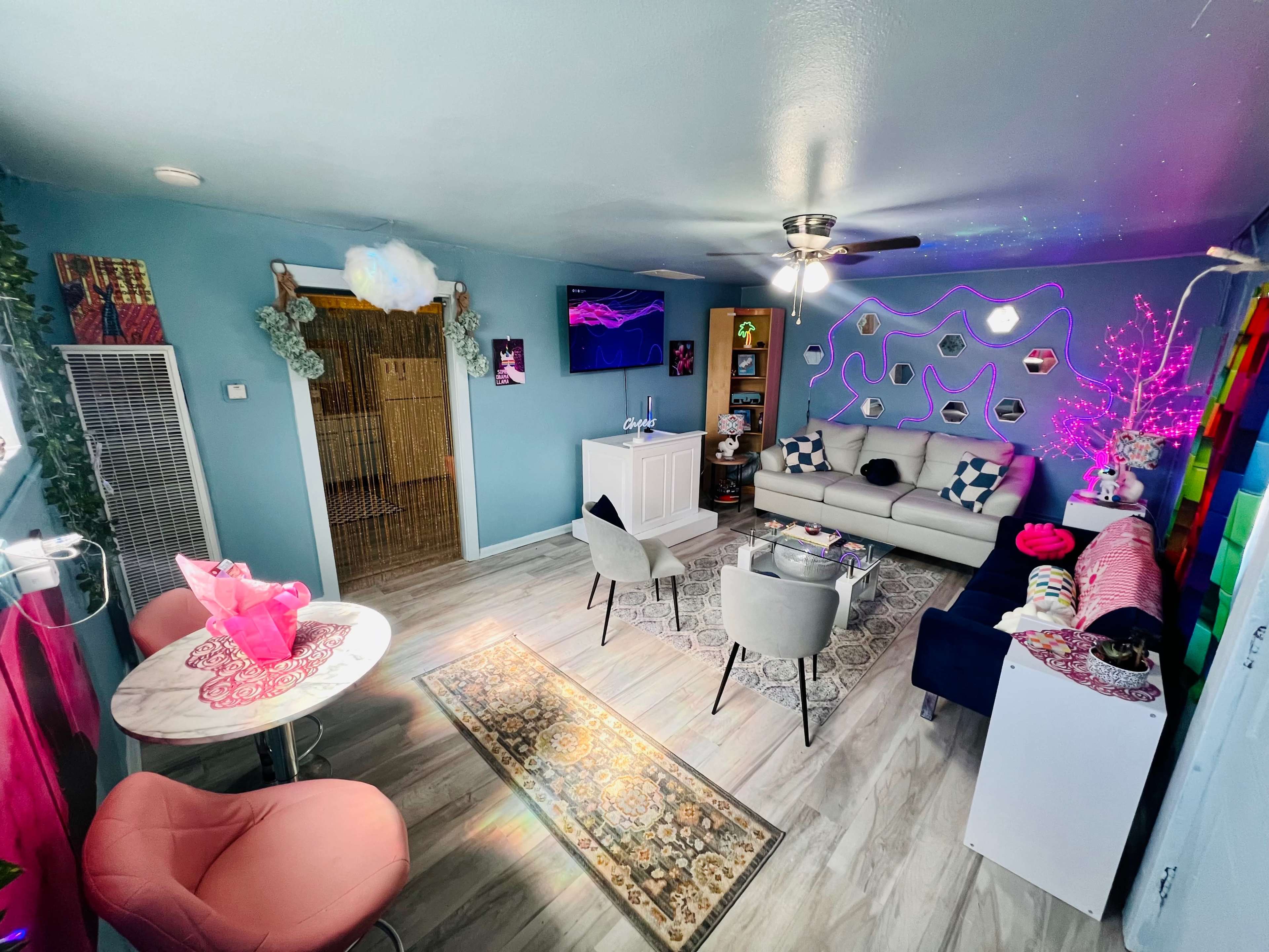 A brightly decorated living room features a light blue color scheme, a gray sofa, a dining table, and various decorative elements like colorful artwork and a TV.