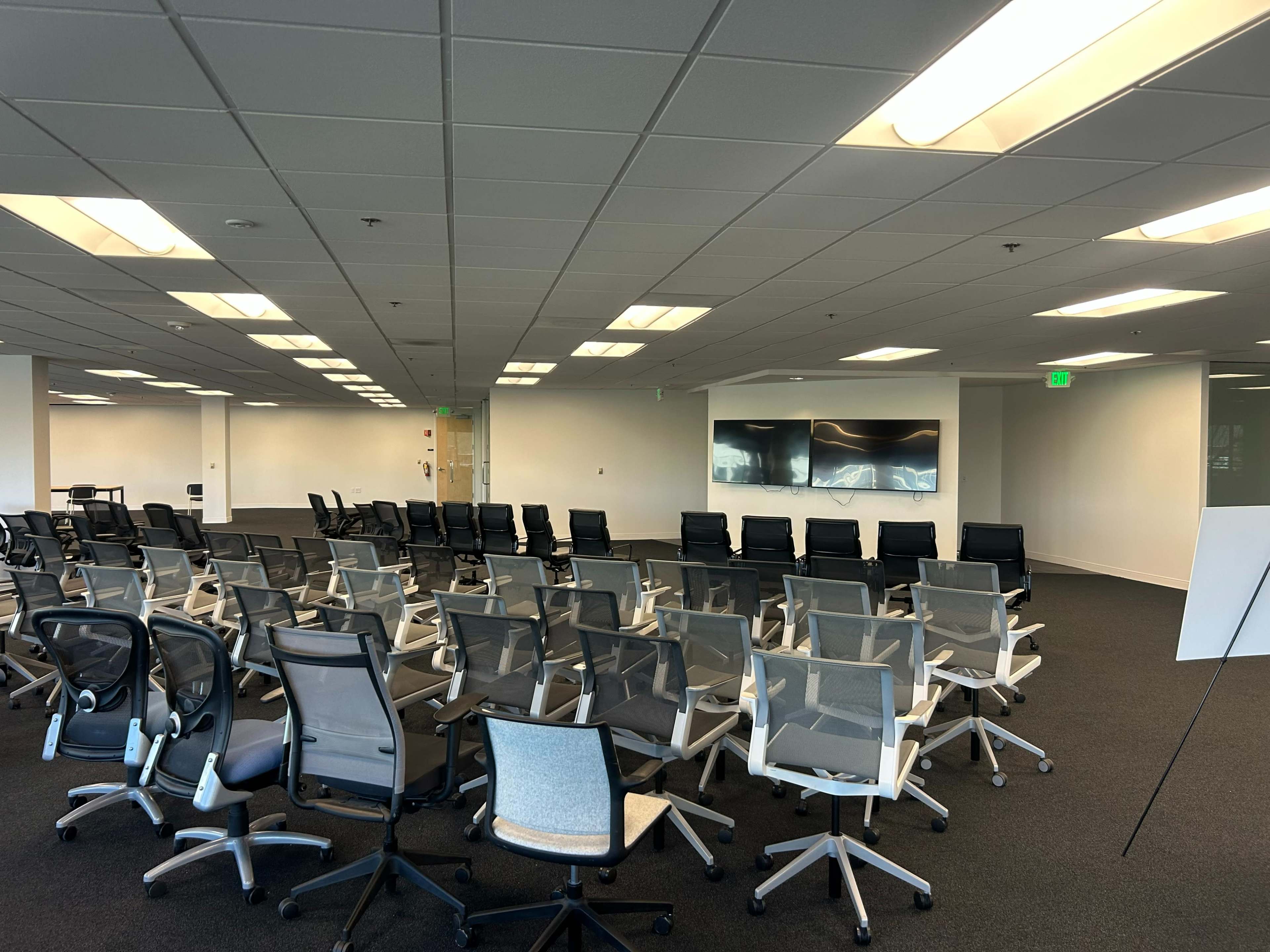 The image shows a modern office space with rows of chairs arranged facing a wall containing three screens, under overhead lighting.