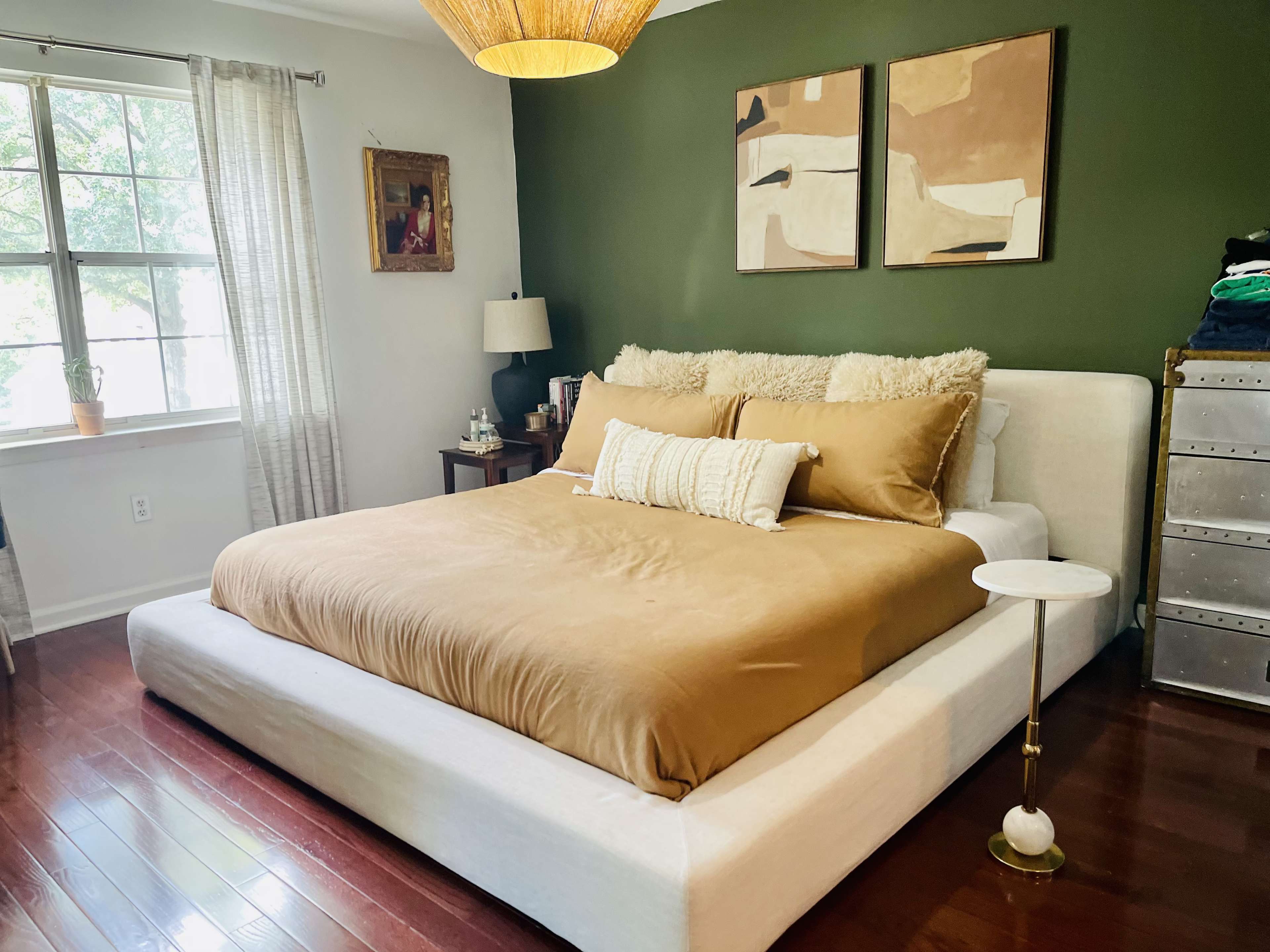 The image shows a bedroom with a large bed featuring a brown duvet, two abstract paintings on a green wall, and a bedside table next to a window.
