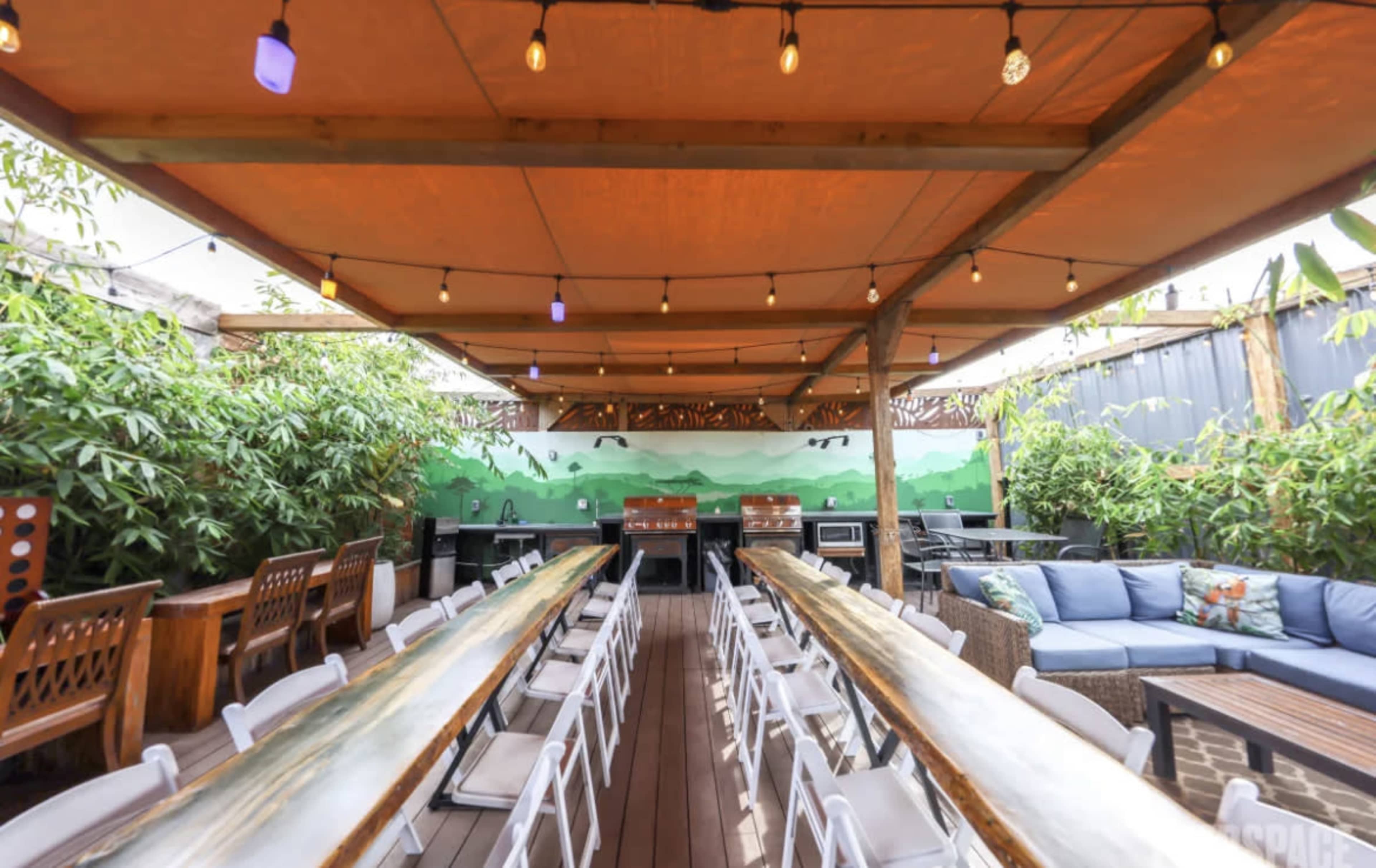The image depicts an outdoor dining area with long tables, chairs, and a seating arrangement under a canopy of lights and a painted mural in the background.