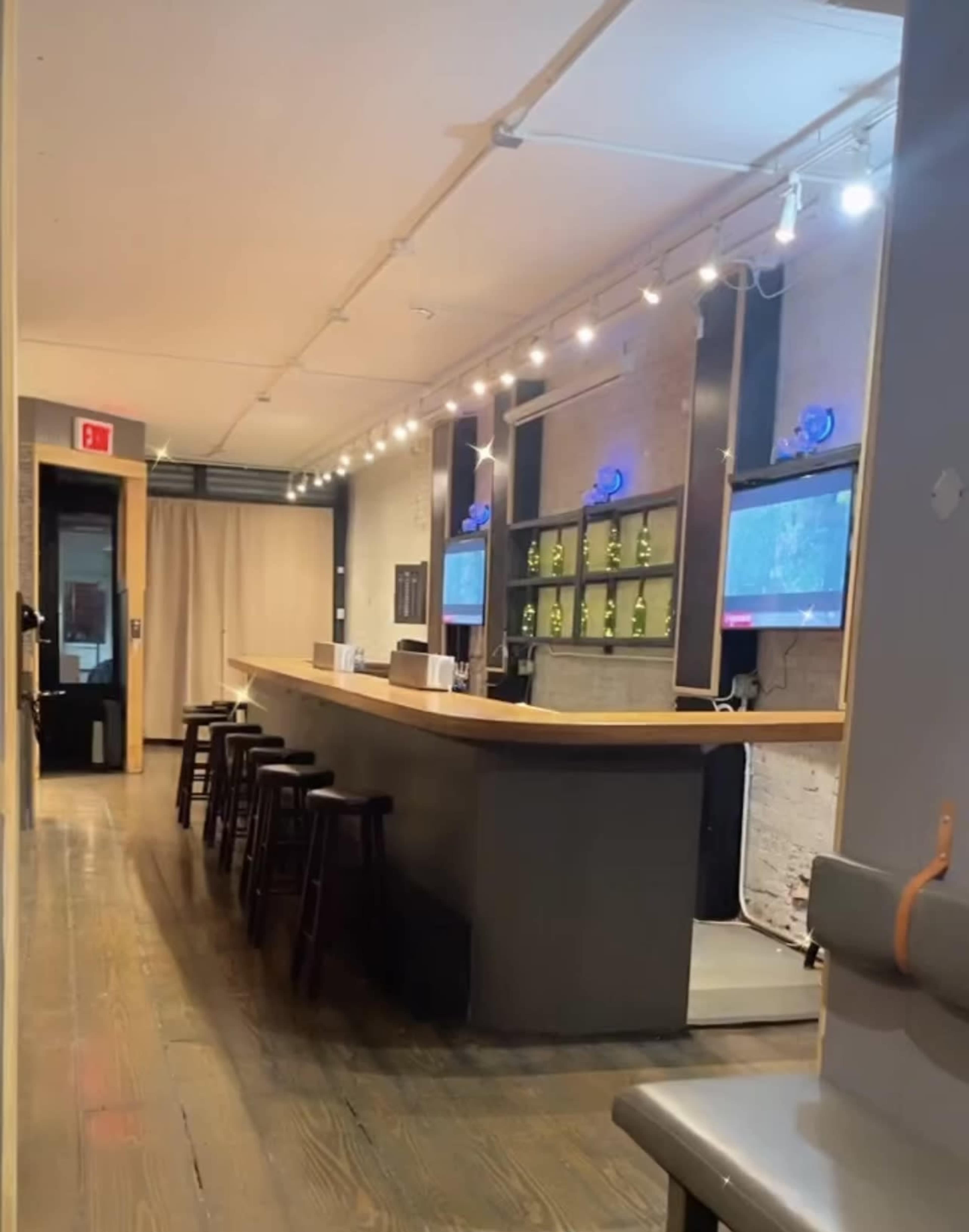 A dimly lit bar area with a long wooden counter, several high stools, and illuminated shelves behind the bar.