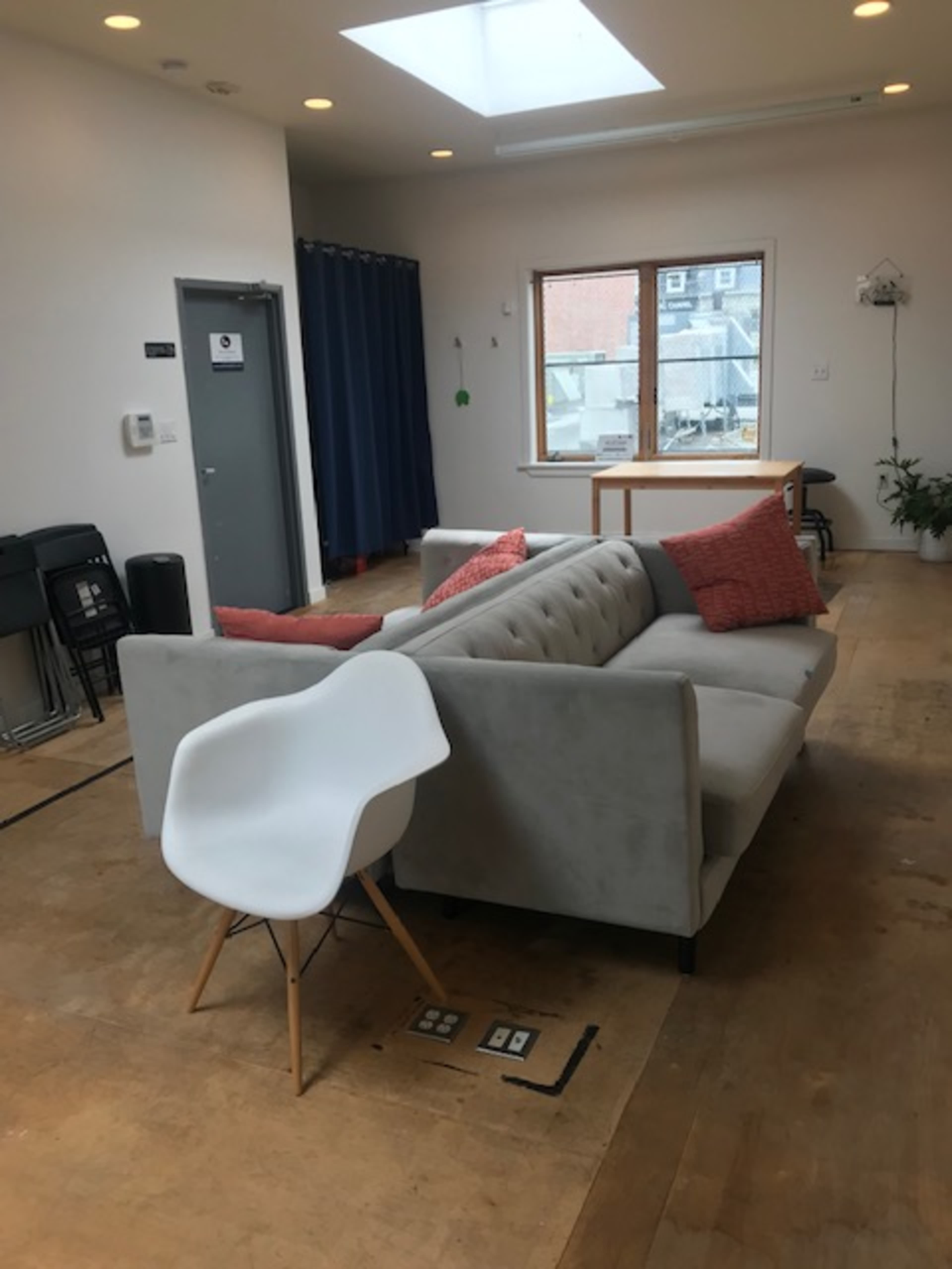 A gray sectional sofa with orange cushions is positioned in a room featuring a large window and a mix of wooden flooring and scattered furniture.