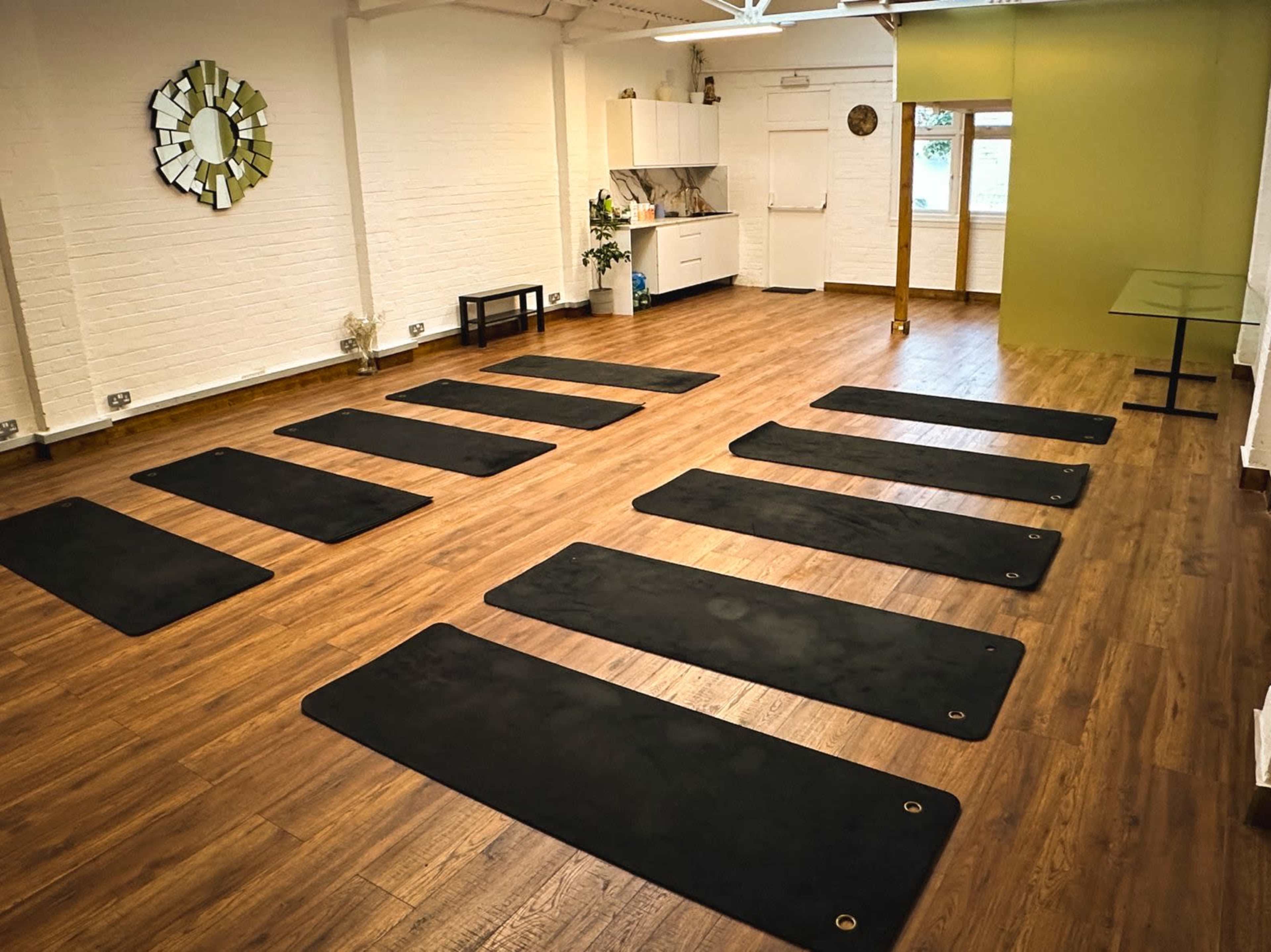 The image shows a spacious yoga studio featuring several black yoga mats arranged neatly on a wooden floor, with green and white walls and minimal furniture.