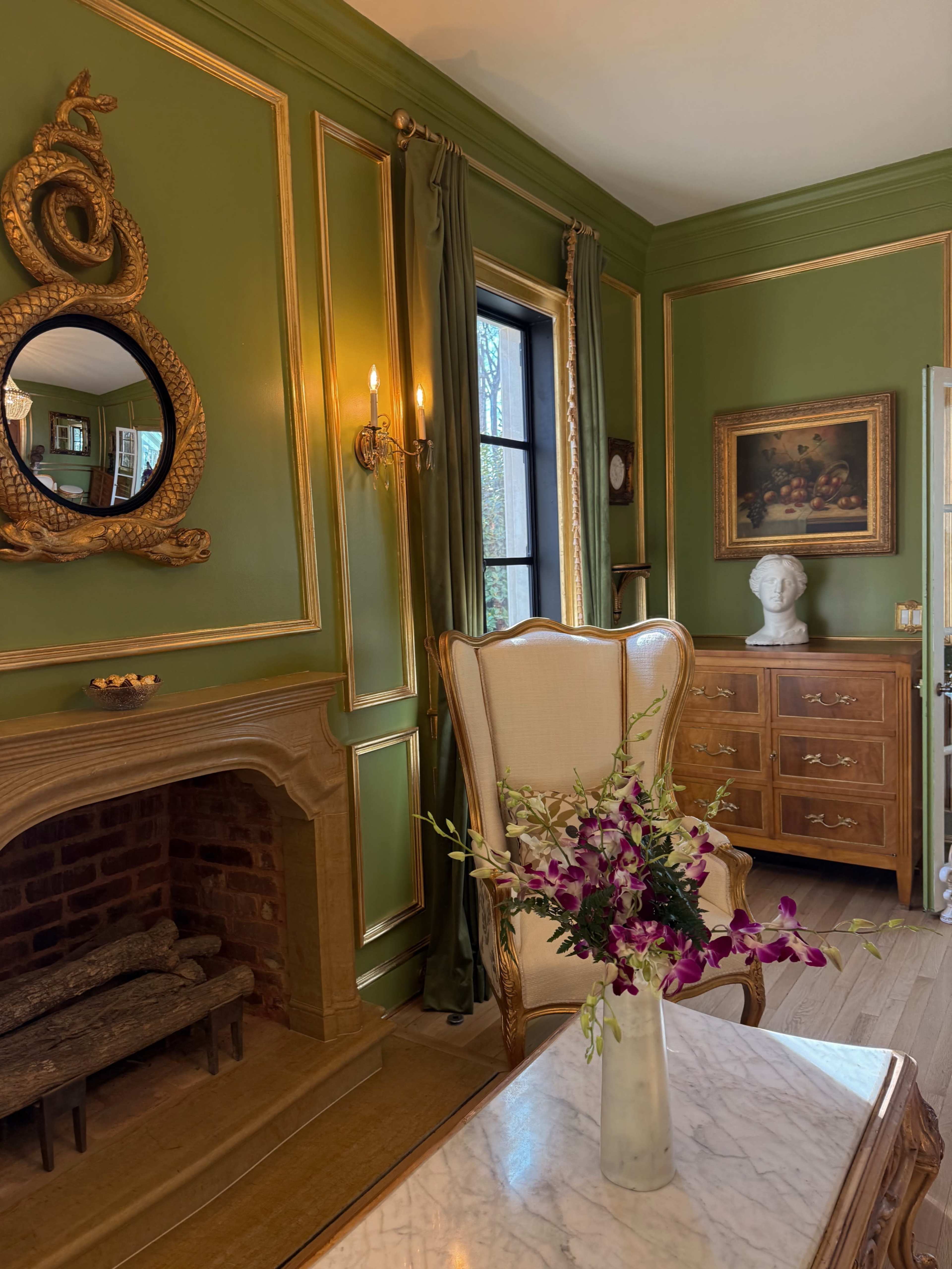 The image features a room with green walls adorned with gold trim, containing a fireplace, a decorative mirror, a bust sculpture, and a wooden dresser, along with a floral arrangement on a marble table.